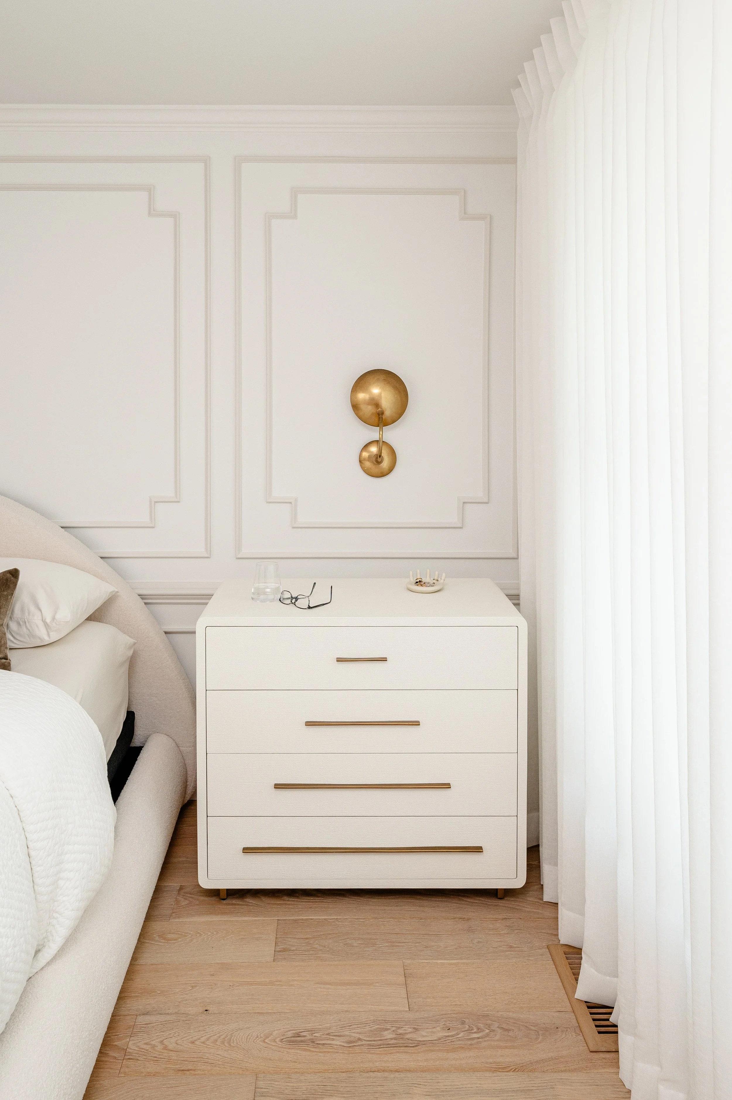 A white bedroom nightstand with five drawers and gold handles, placed next to a bed and in front of white curtains, with a gold wall sconce above it, on a light wood floor.