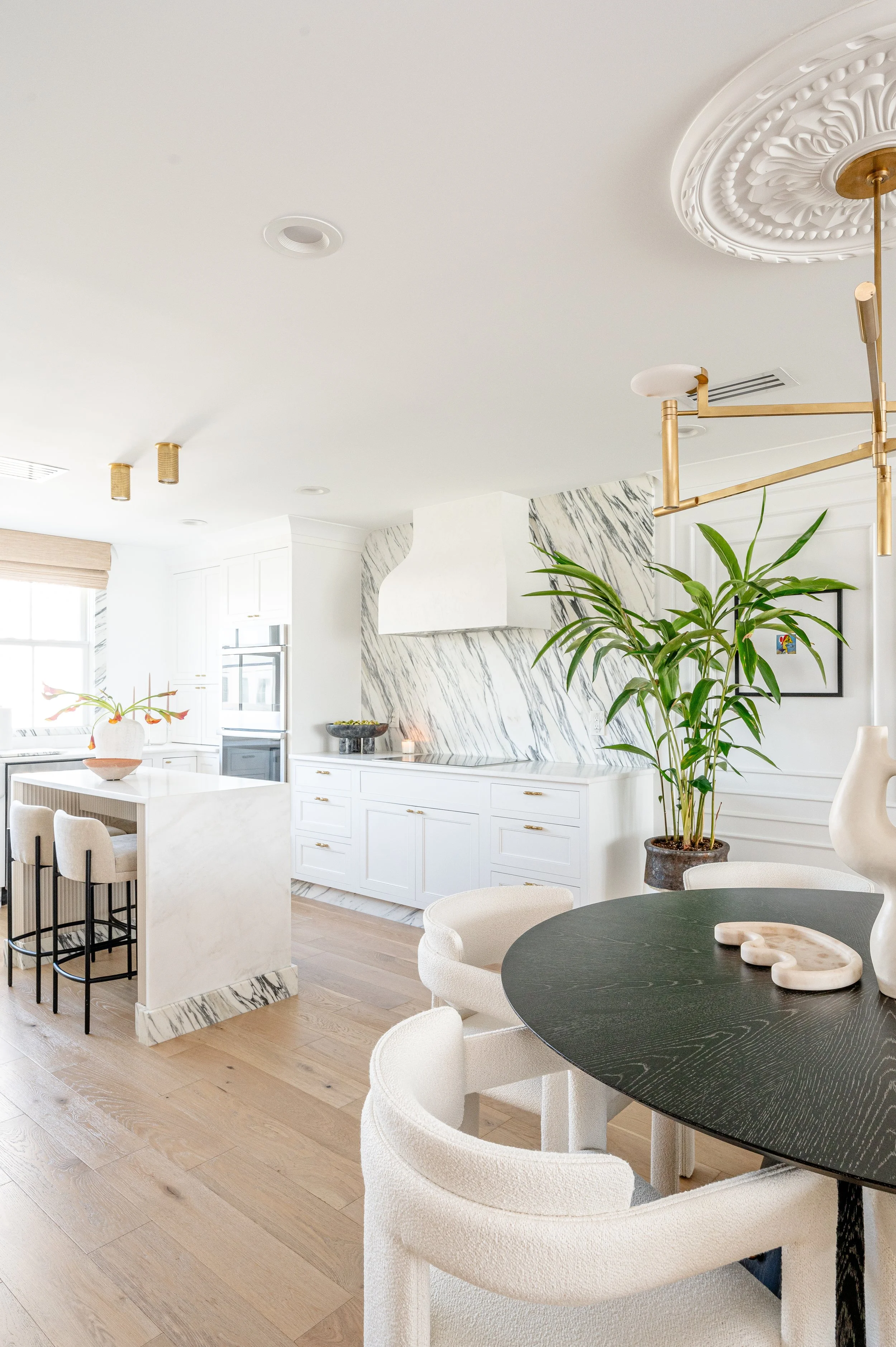 A bright, modern kitchen and dining area with white cabinets, a marble backsplash, a black round dining table with cream chairs, and a large plant in a pot. There's a marble kitchen island and a window with blinds.