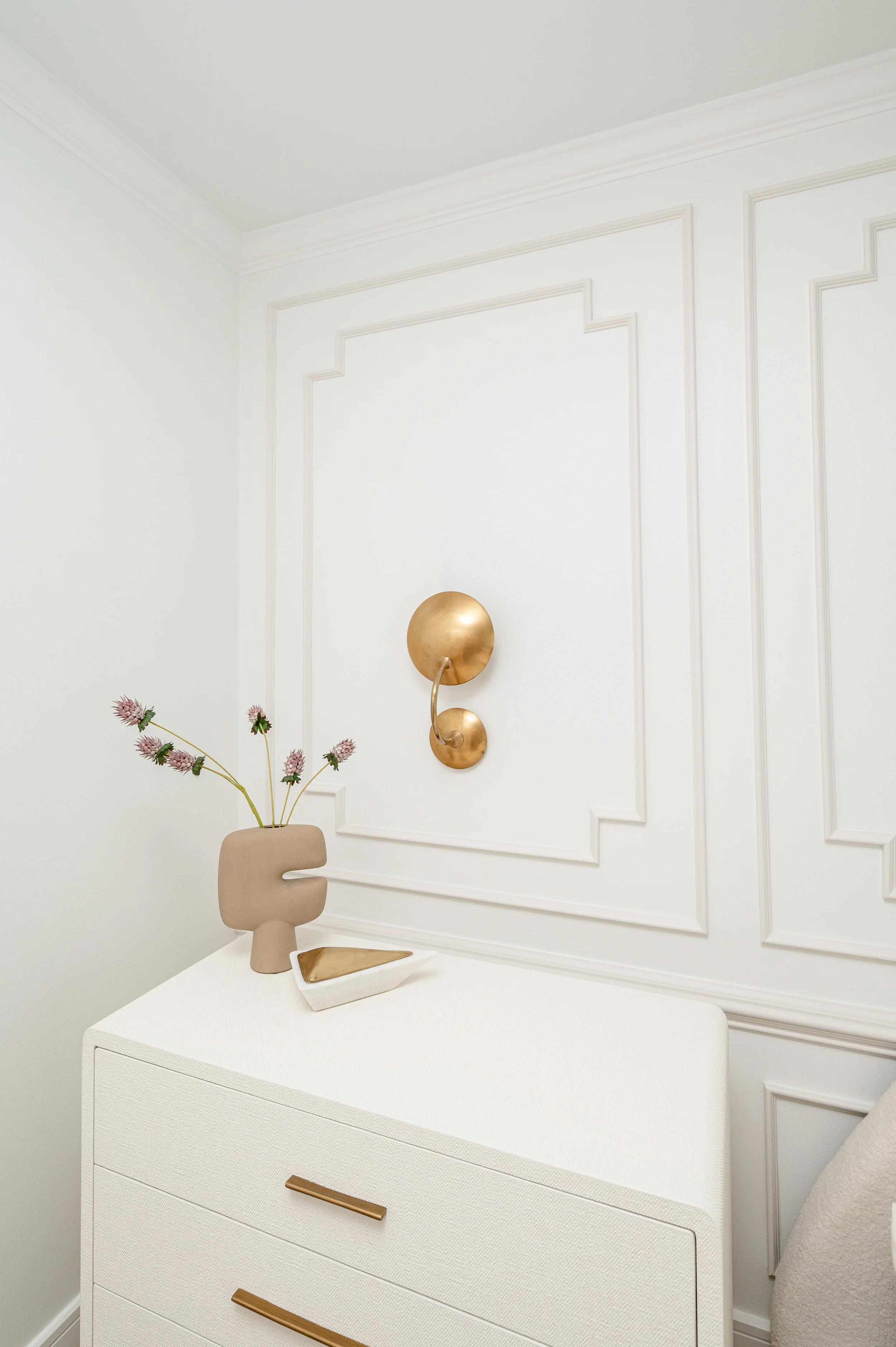 Decorative interior featuring a white dresser, a beige vase with purple flowers, a geometric tray, and a gold wall sculpture on a white paneled wall.