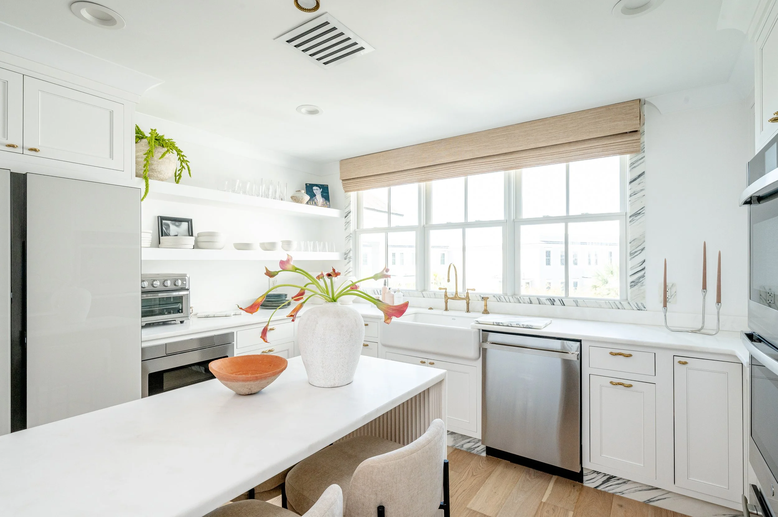 Bright kitchen with white cabinetry, marble countertops, and a large window. A white island with a large white vase with pink flowers, a brown bowl, and beige chairs. Gold fixtures, a stainless steel dishwasher, and decorative items on open shelves.
