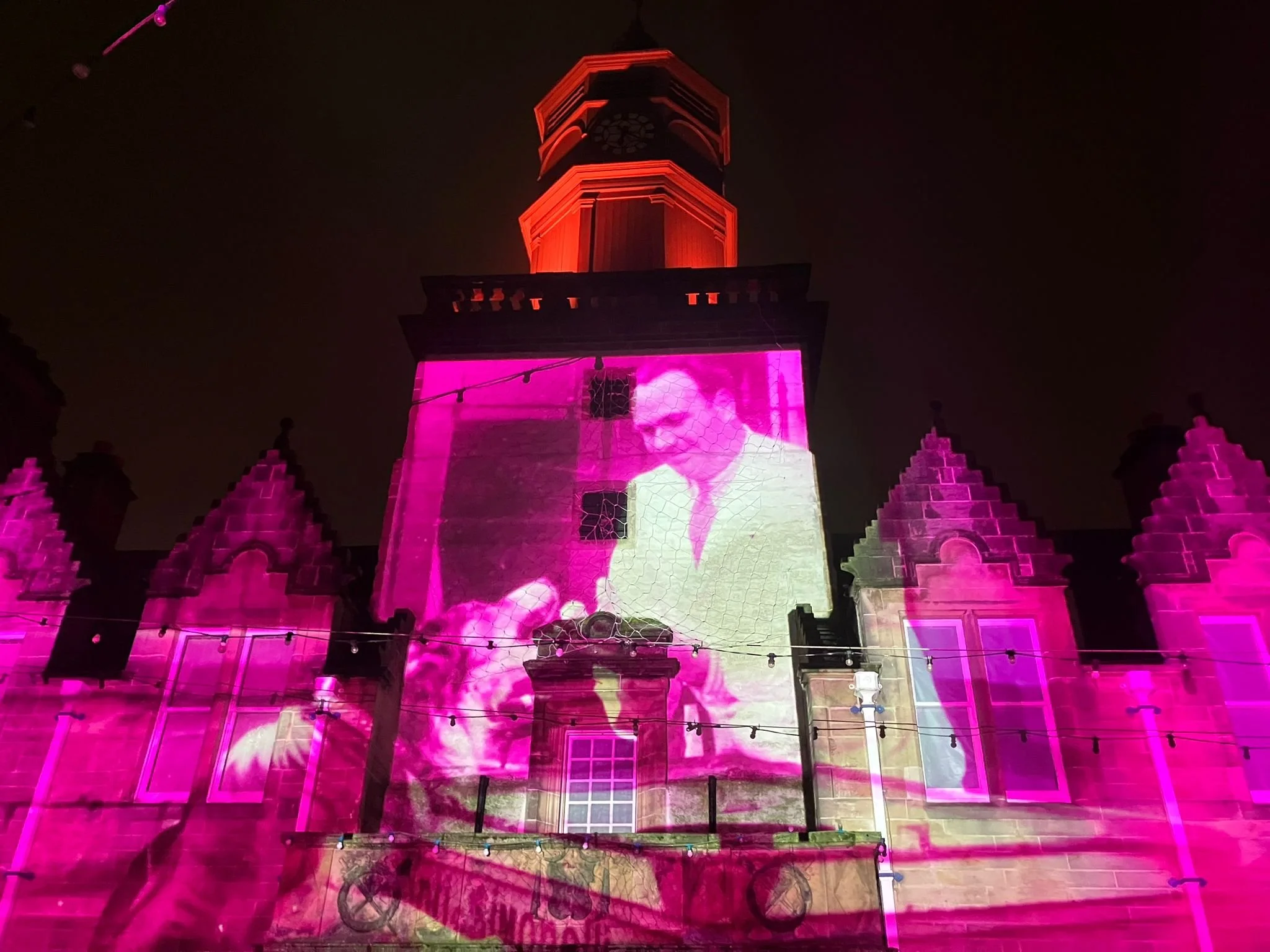 Nighttime view of a tall historic building illuminated with pink lights and projection, showing a woman's face on the facade, topped with a clock tower.