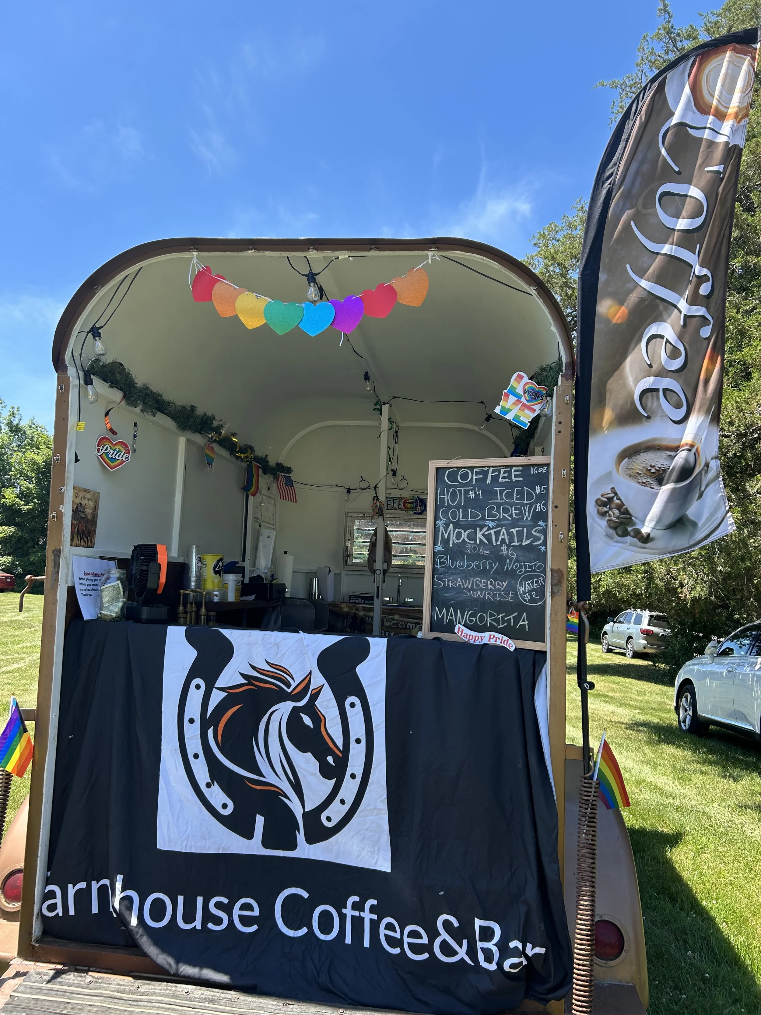 A food truck with a black banner displaying white text for a coffee and bar business. The truck is decorated with rainbow-colored heart and pride flags, and a black and white logo of a horse's head surrounded by a circle of hoofprints. Inside the tru