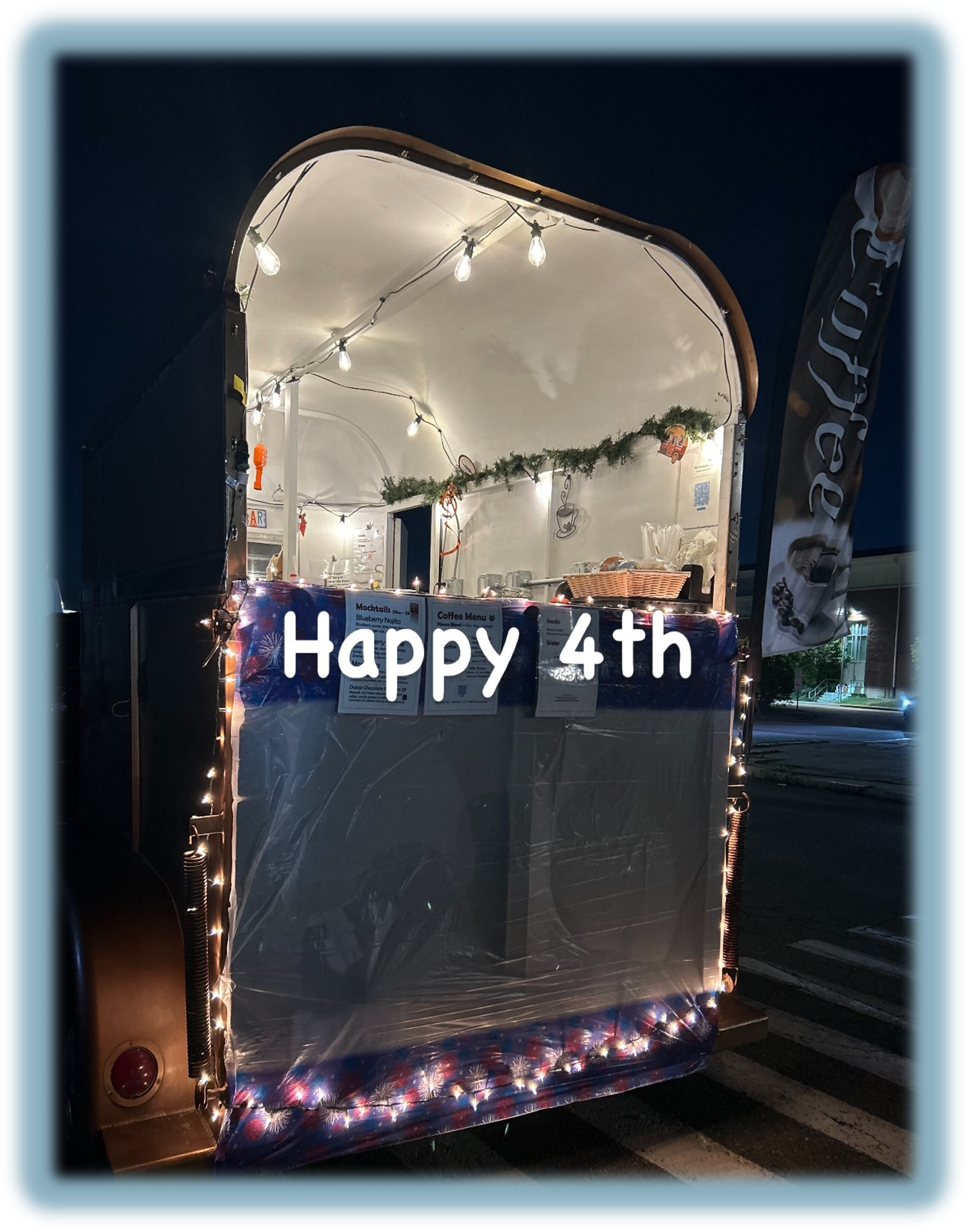 A mobile food stand decorated with white string lights and holiday decorations, celebrating the 4th of July, with a banner that reads 'Happy 4th'.