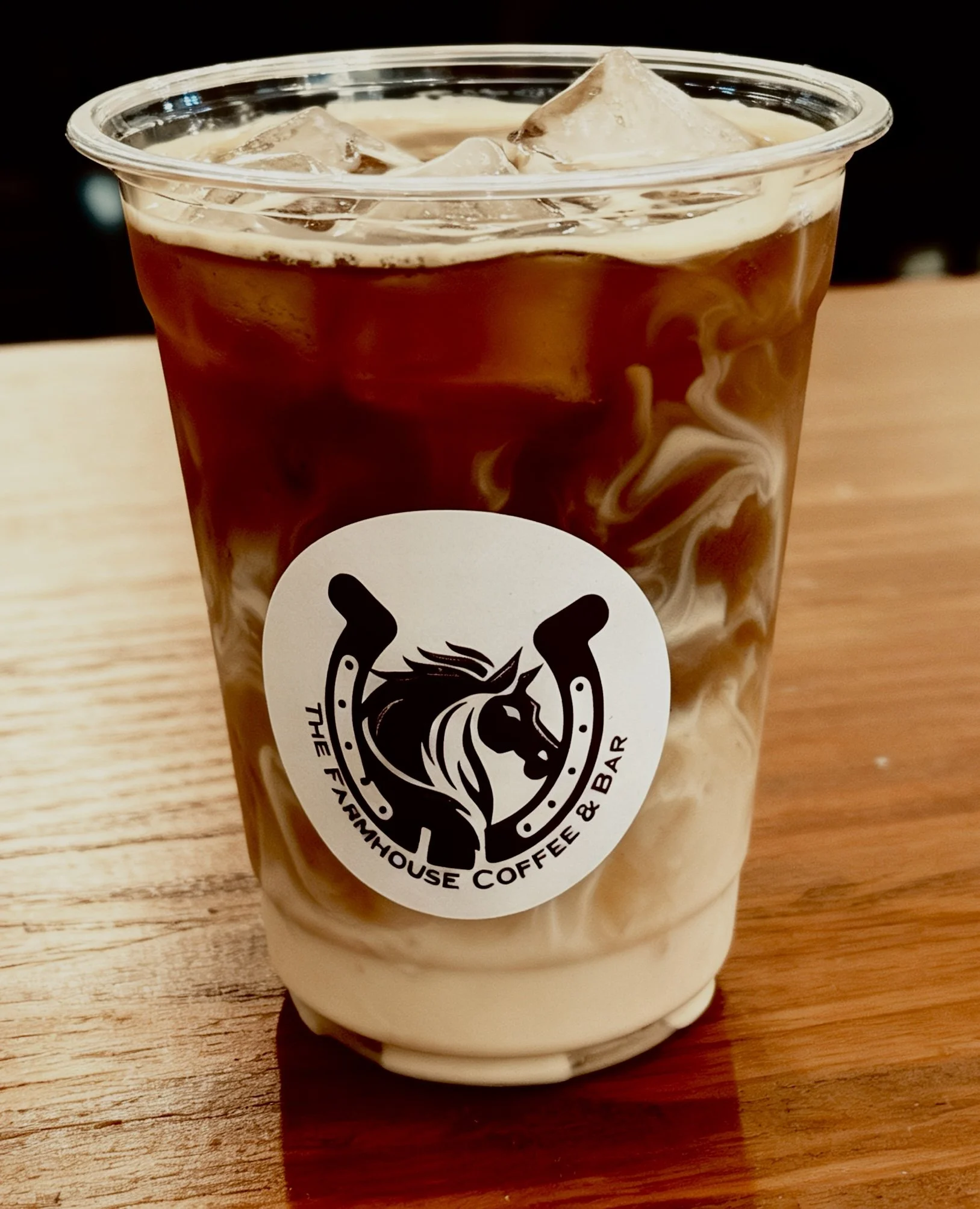 A clear plastic cup filled with iced coffee, swirled with cream, placed on a wooden surface, featuring a black and white logo of a horse's head.