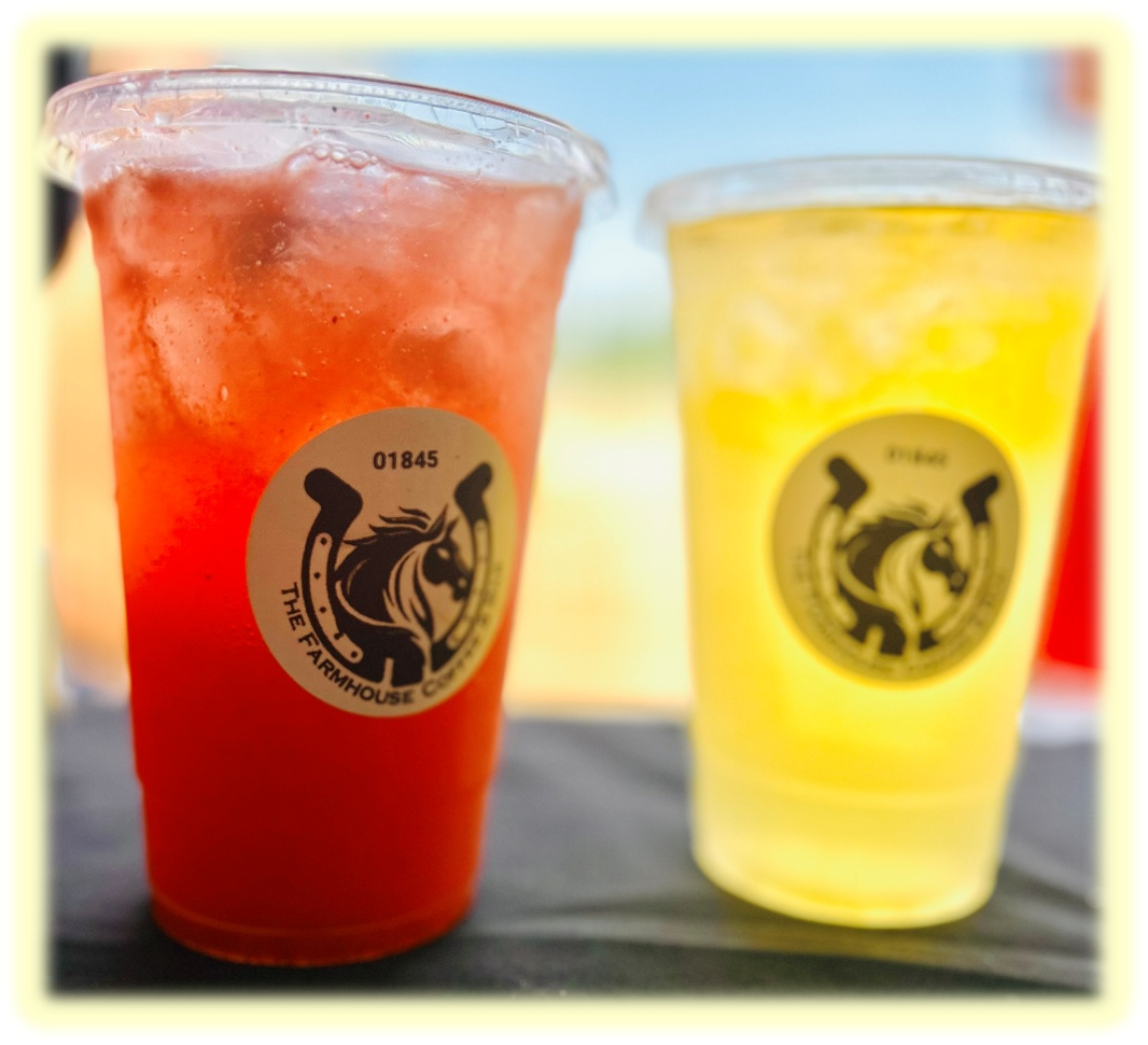 Two cups of cold beverages, one red and one yellow, on a table with a logo of a horse's head enclosed in a horseshoe from The Farmhouse. The red drink appears to be a slush or iced beverage, and the yellow drink looks like a lemonade or citrus soda.