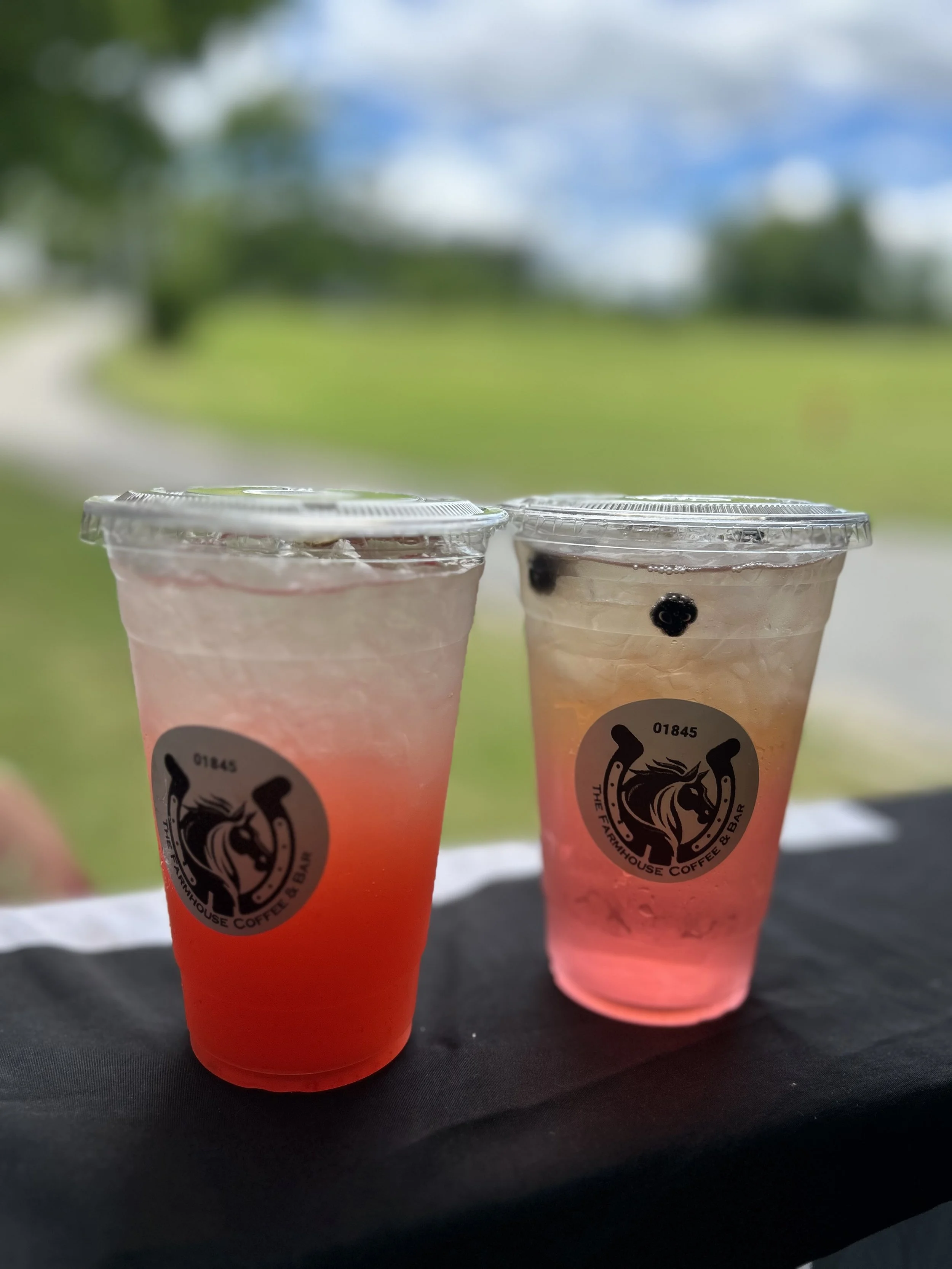 Two cups of pink and orange drinks with clear plastic lids and black logos, placed on a dark surface outdoors near a grassy area with trees and sky in the background.