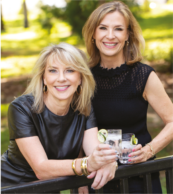 Two women smiling outdoors, holding glasses of clear beverages with lime slices, standing behind a black railing, with green trees and sunlight in the background.