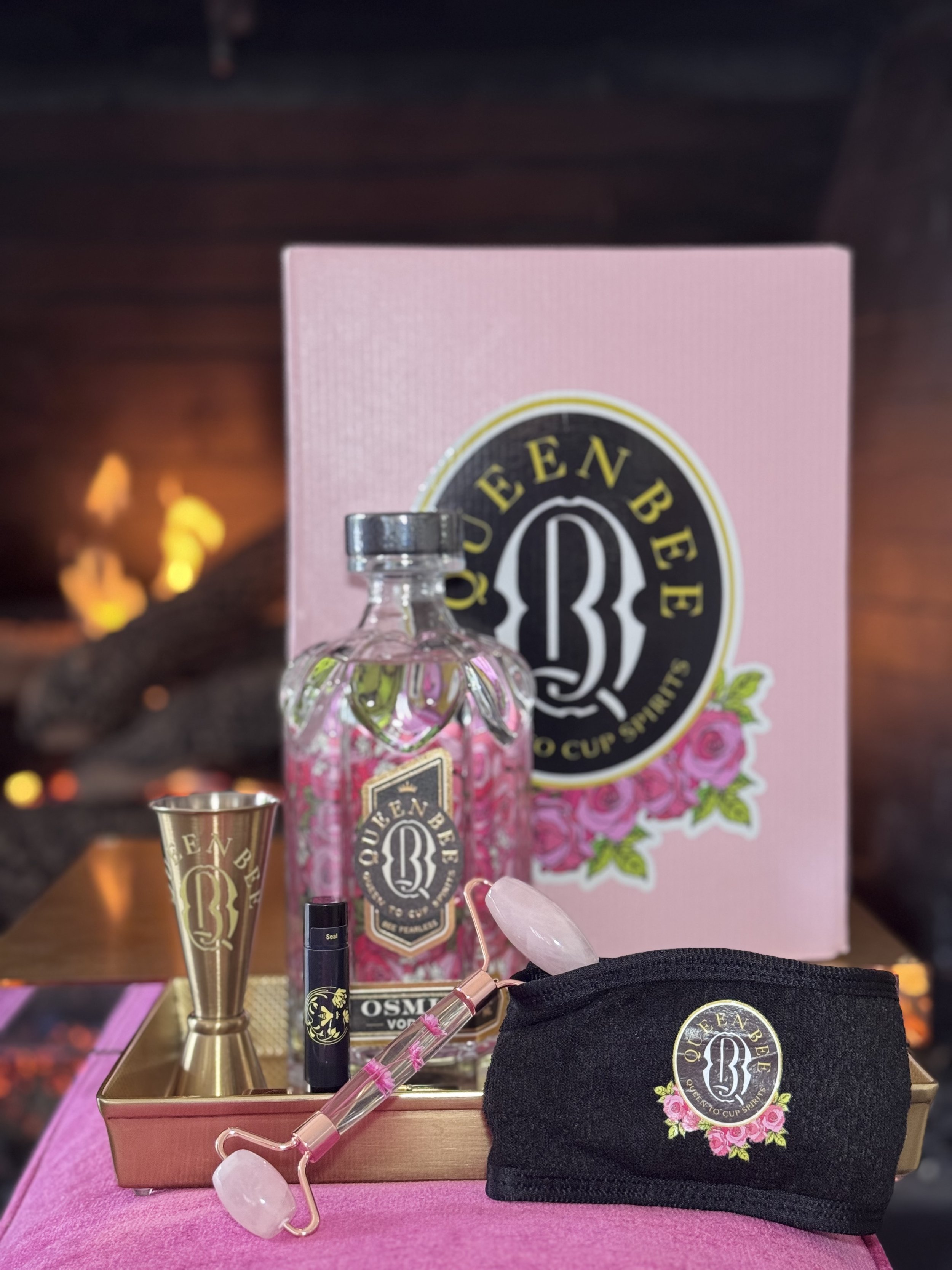 A beauty set displayed on a pink surface with a background of a fireplace and a log. The set includes a decorated glass bottle, a gold shot glass, a black tube, a pink facial roller, a pink gua sha, and a black pouch, all branded with a QueenBee logo