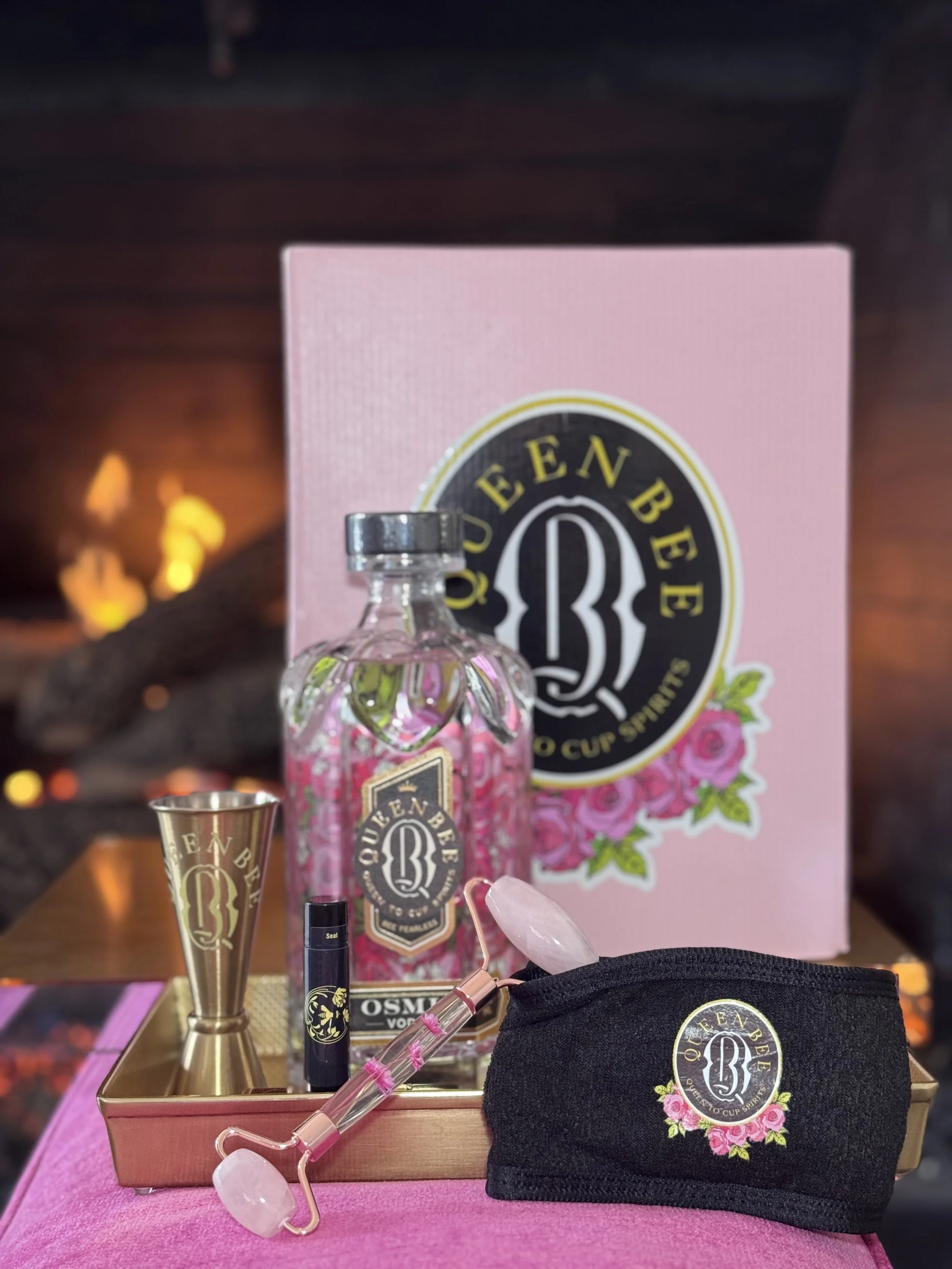 A pink QueenBee vodka bottle and related accessories on a tray, including a shot glass, a black pouch, a facial roller, a lip balm, and a pink rose-themed box in the background.