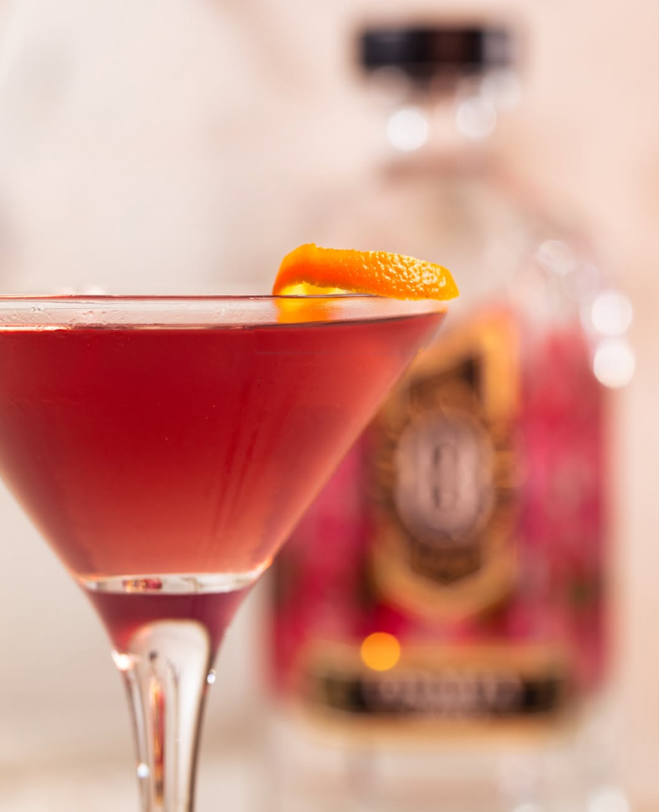 A cocktail glass filled with a red drink, garnished with an orange peel, with a blurred bottle in the background.