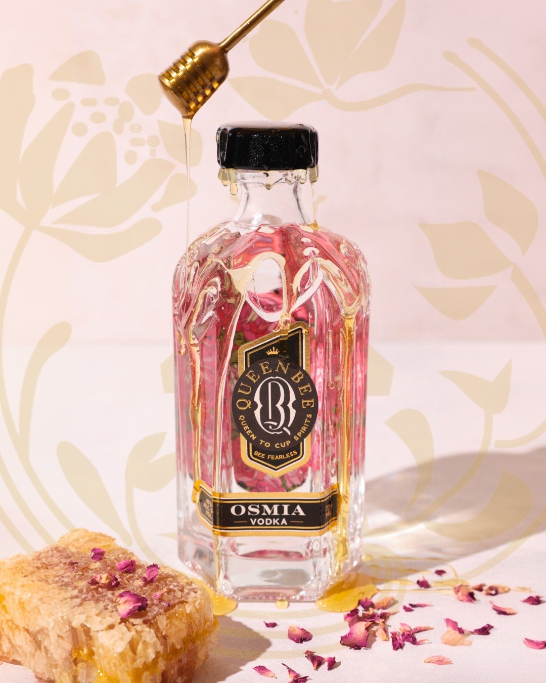 A bottle of Queen Bee Osma Vodka with honey dripping from a honey dipper above it. A honeycomb and rose petals are on a white surface in front of the bottle, with a beige floral pattern background.