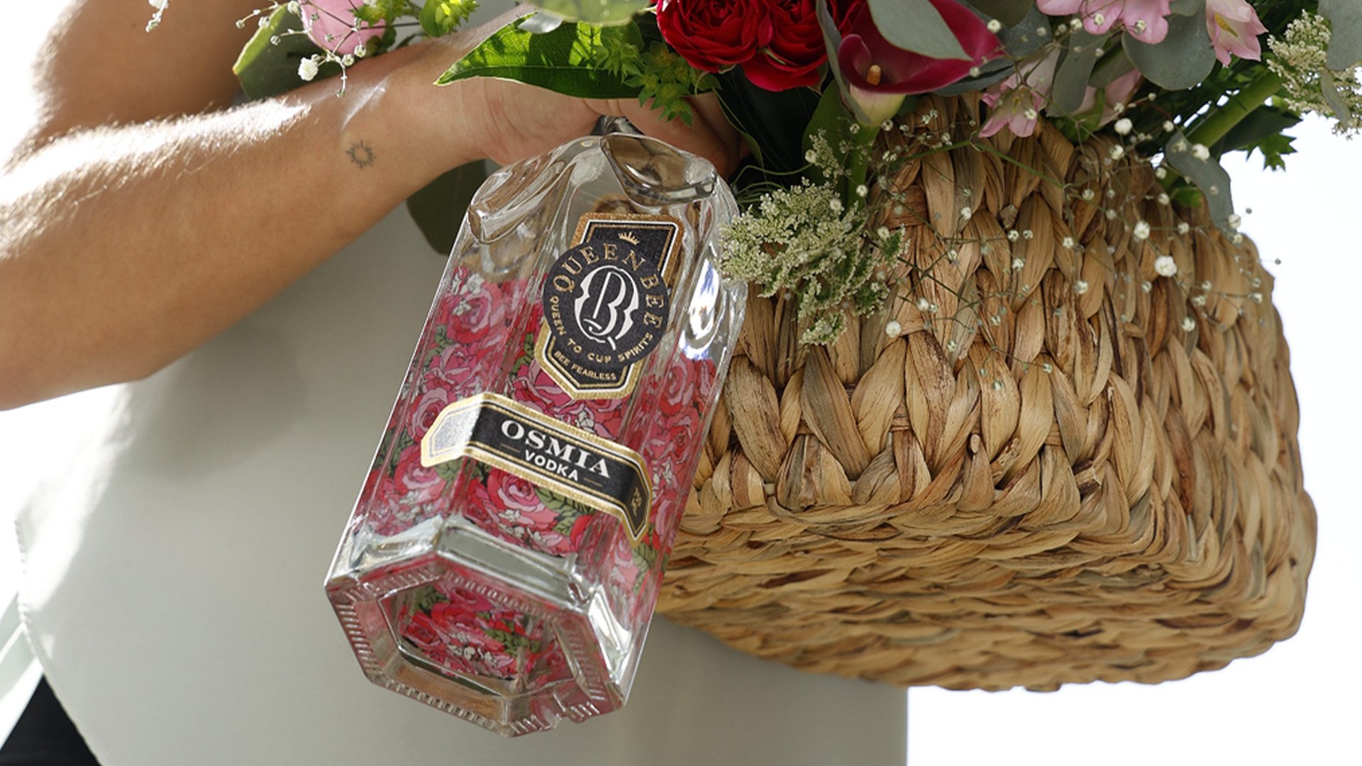 A hand holding a Queen Bee Amsterdam vodka bottle next to a large woven basket filled with pink, red, and white flowers and greenery.