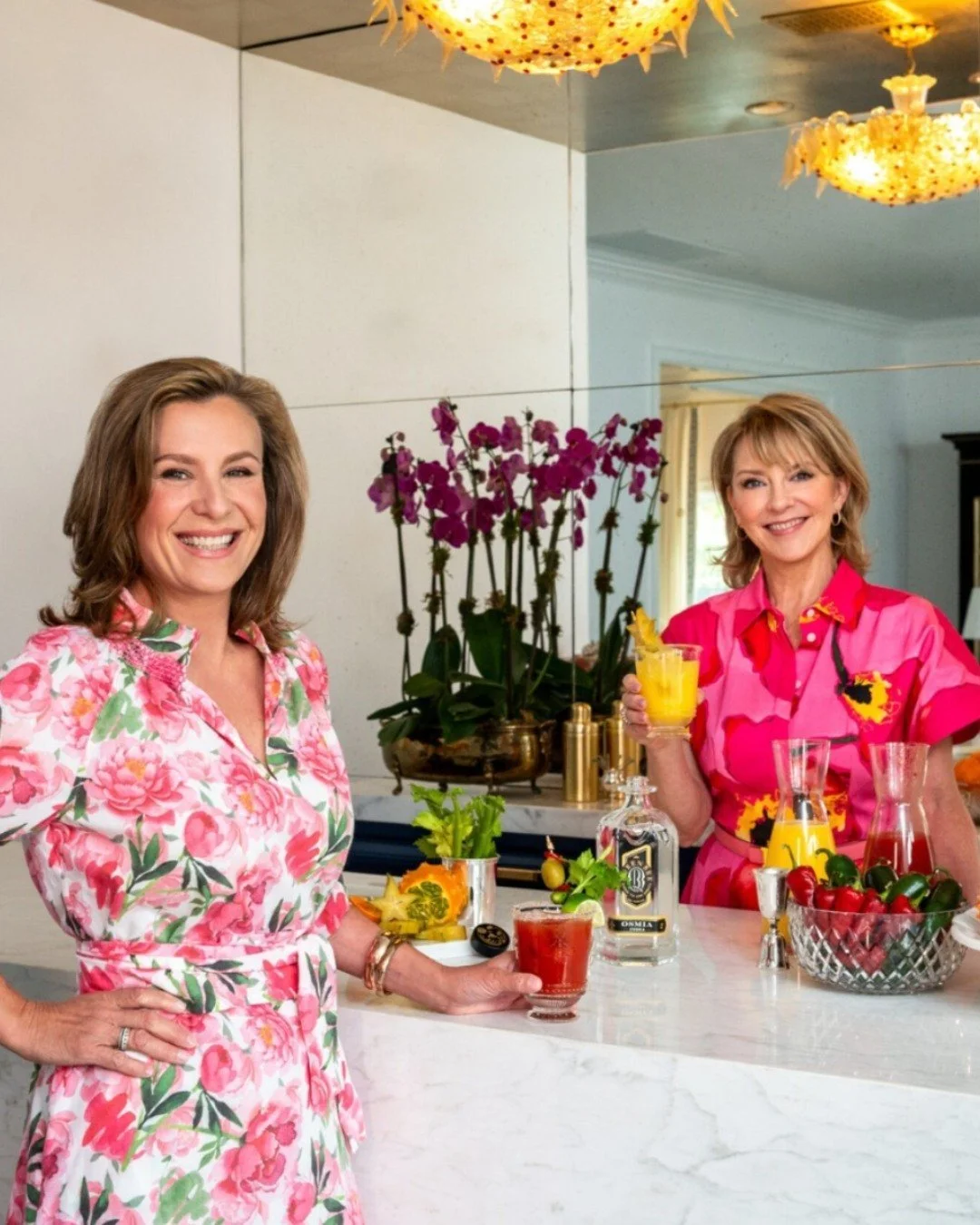 Two women smiling and holding drinks in a bright kitchen with colorful floral attire, a fruit bowl, alcohol bottles, and an orchid plant in the background.