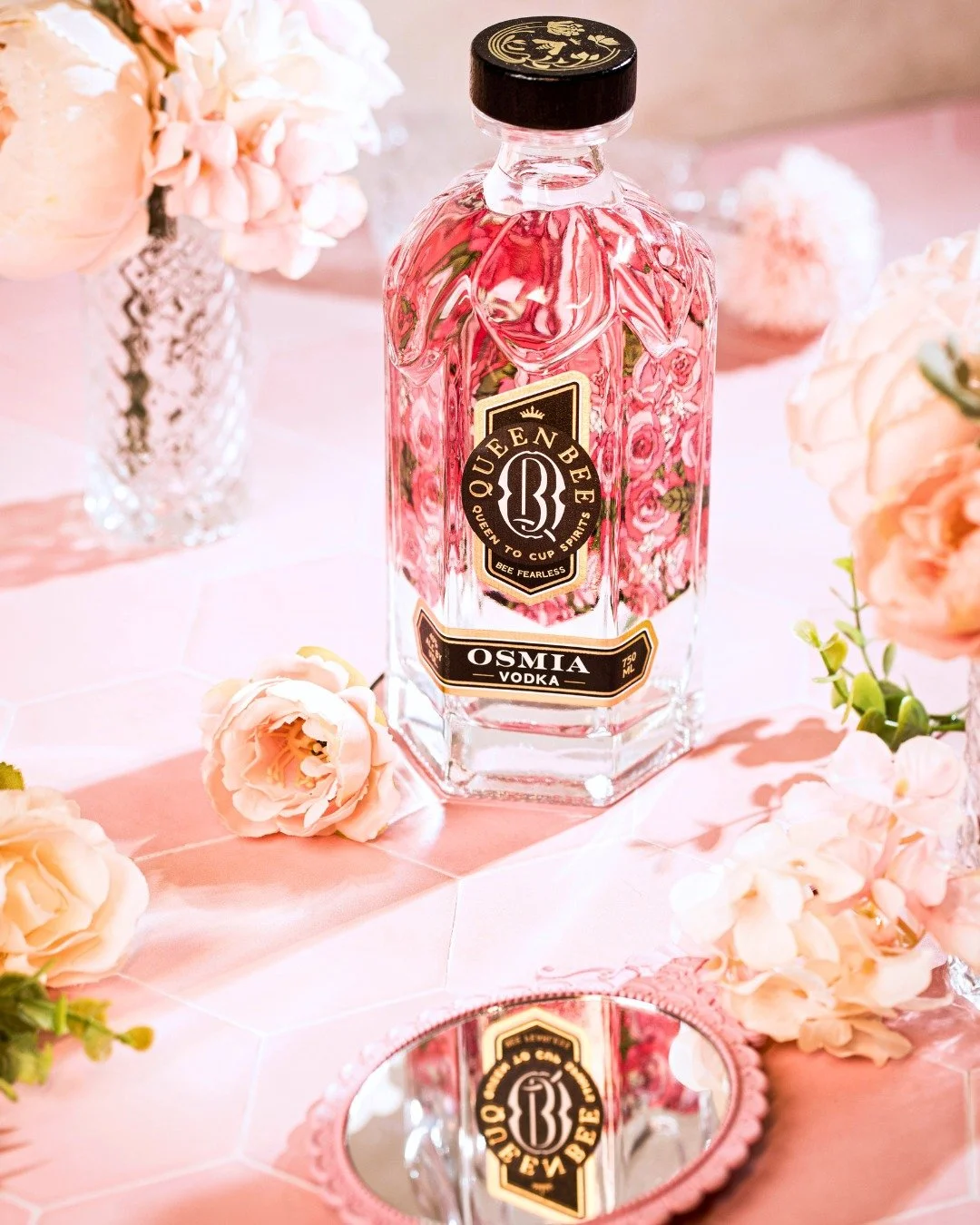 A clear glass bottle of Queen Bee Osmi vodka with pink liquid inside, placed on a pink surface with pink flowers and a small mirror reflecting the bottle.