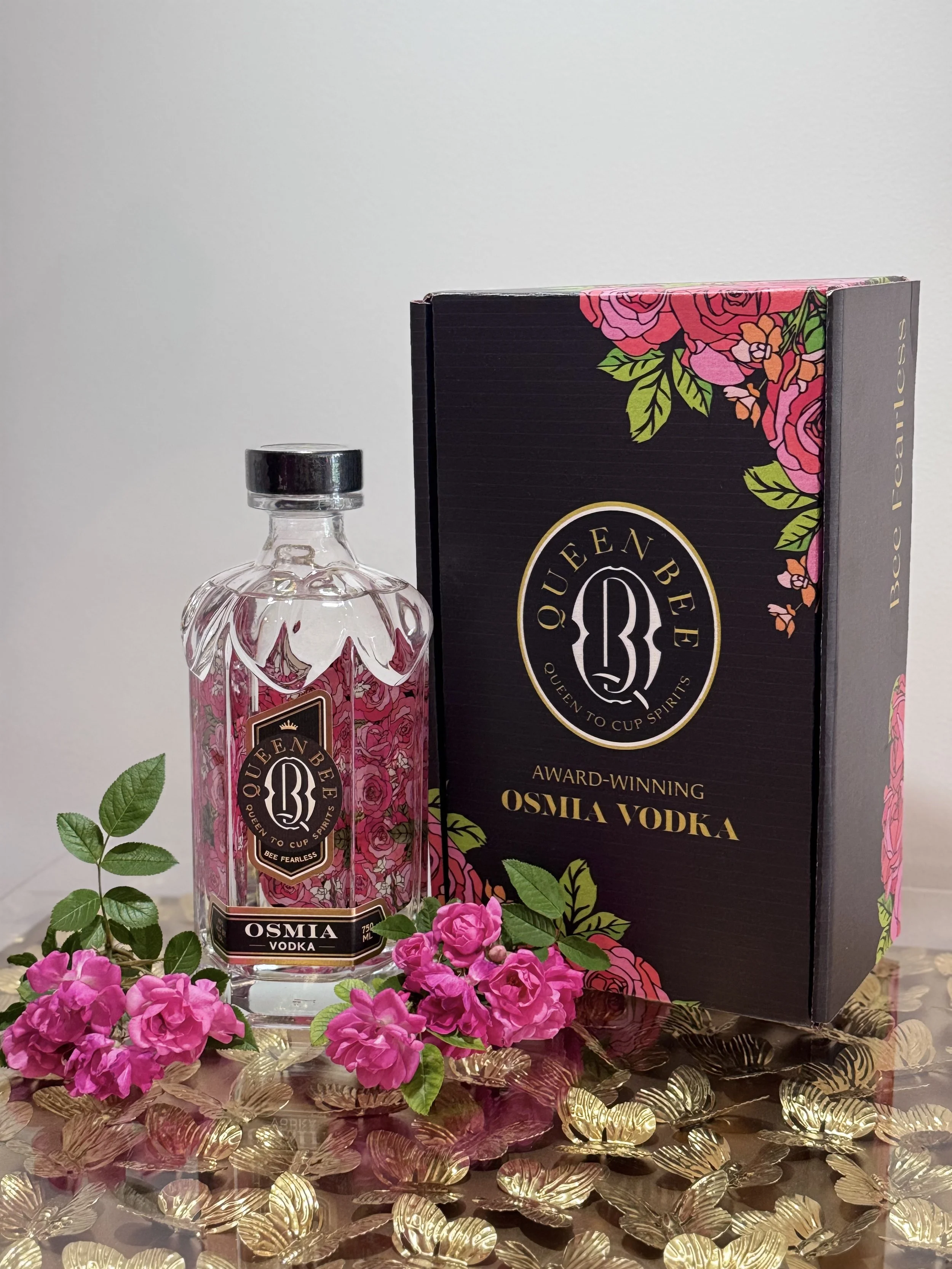 A bottle of Queen Bee Osma Vodka with pink floral label, a black box with pink flowers and gold lettering, pink flowers, and gold decorative items on a reflective surface.