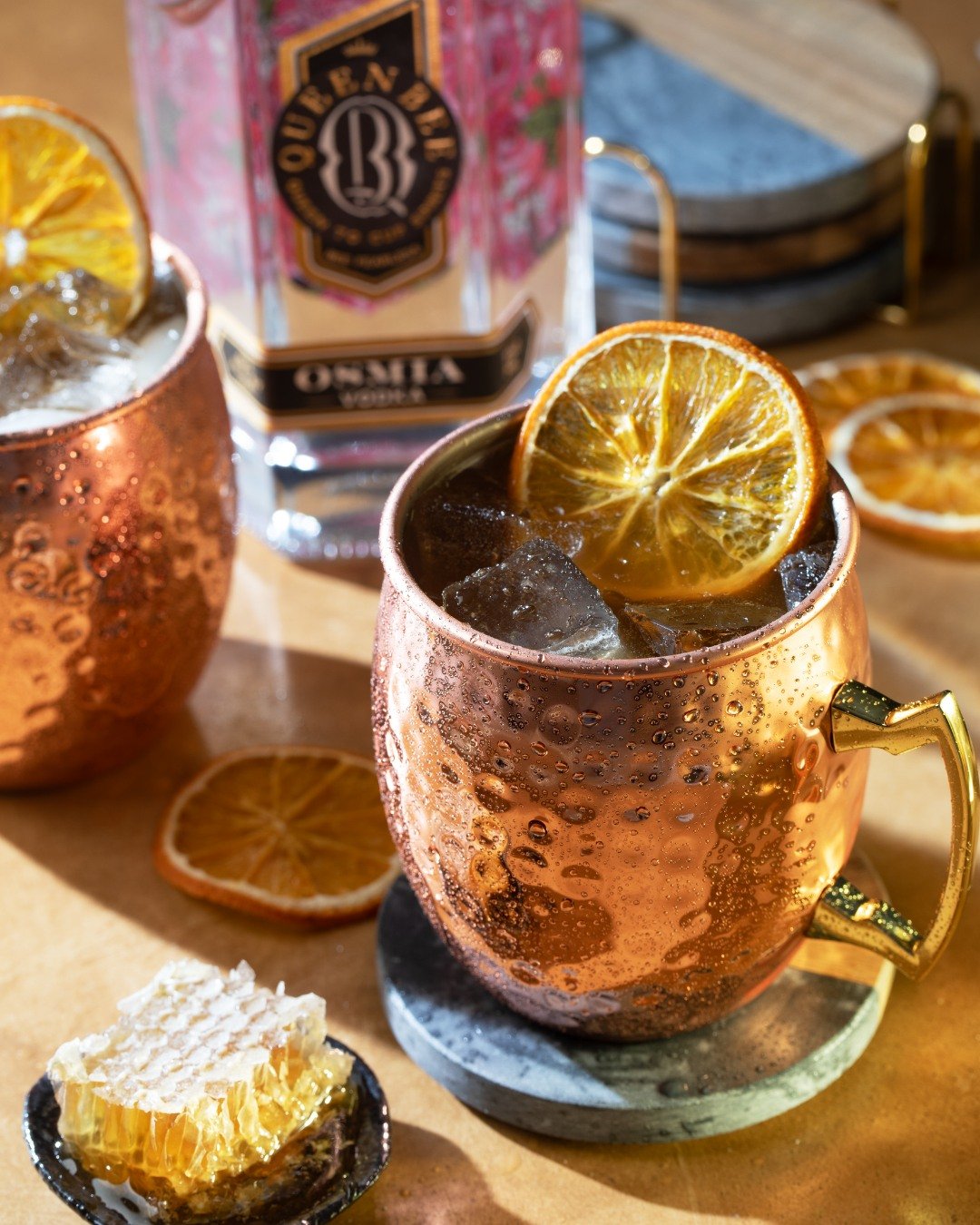 Copper mug with a lemon slice and dark beverage with ice, surrounded by dried lemon slices, a honeycomb, and a packet of Quernby Osina whiskey in the background.