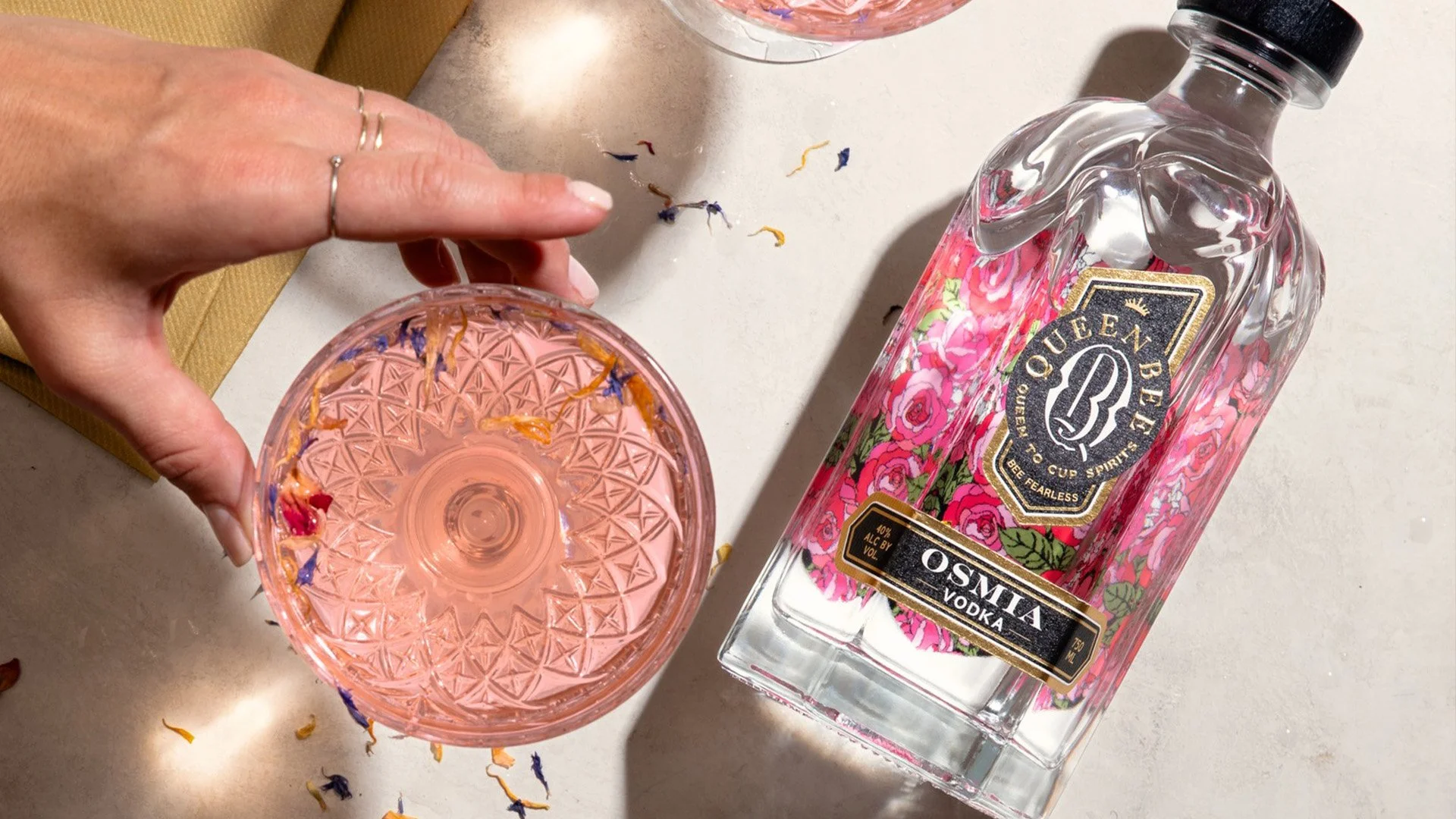 A hand holding a pinkish glass with dried flower petals inside, next to a bottle of Queen Bee Osmia vodka with a floral label. There are dried flower petals scattered on a white countertop.