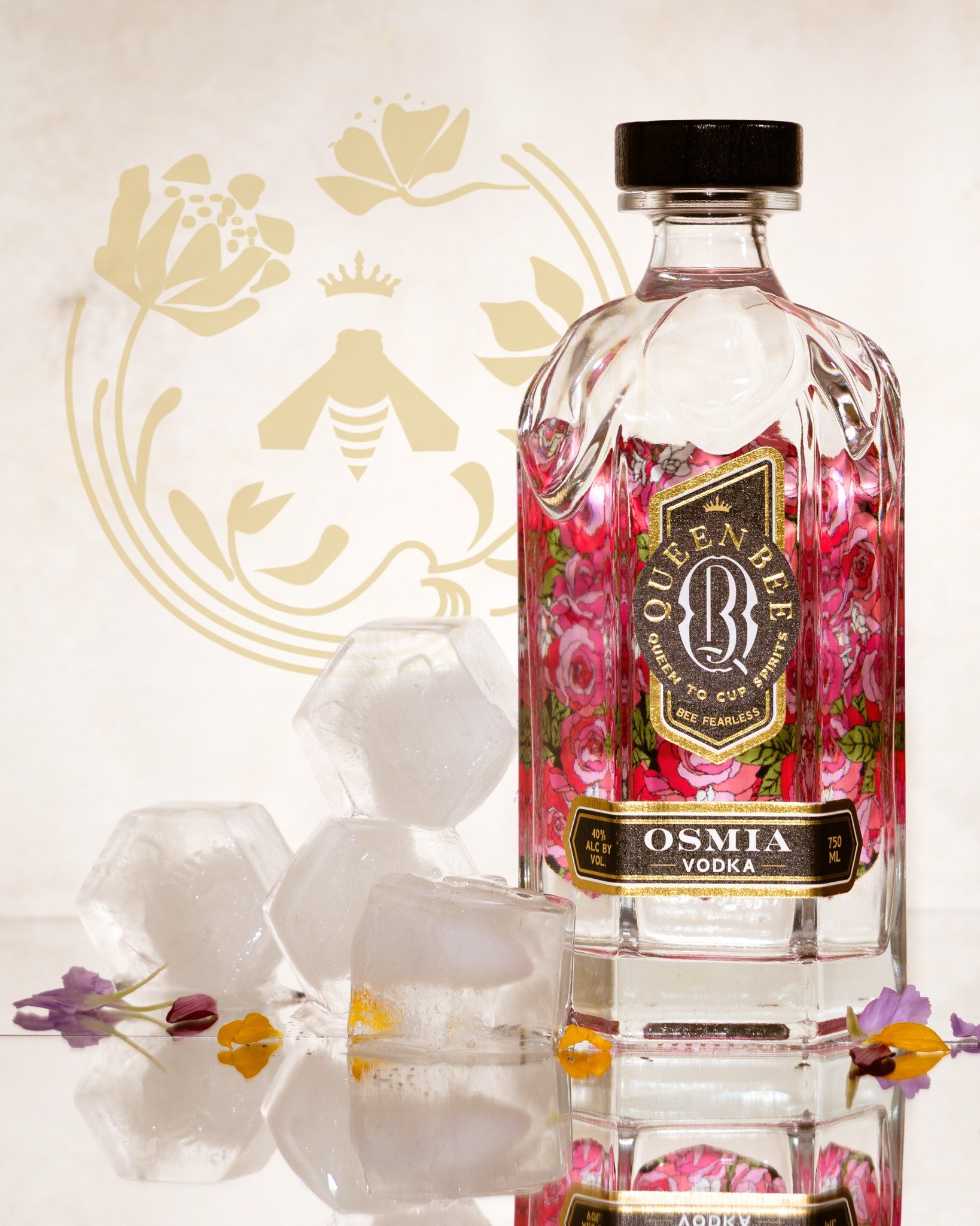 A clear glass bottle of Queen Bee Queen to Cup vodka with pink floral design and a gold label, surrounded by ice cubes and small flower petals on a reflective surface.