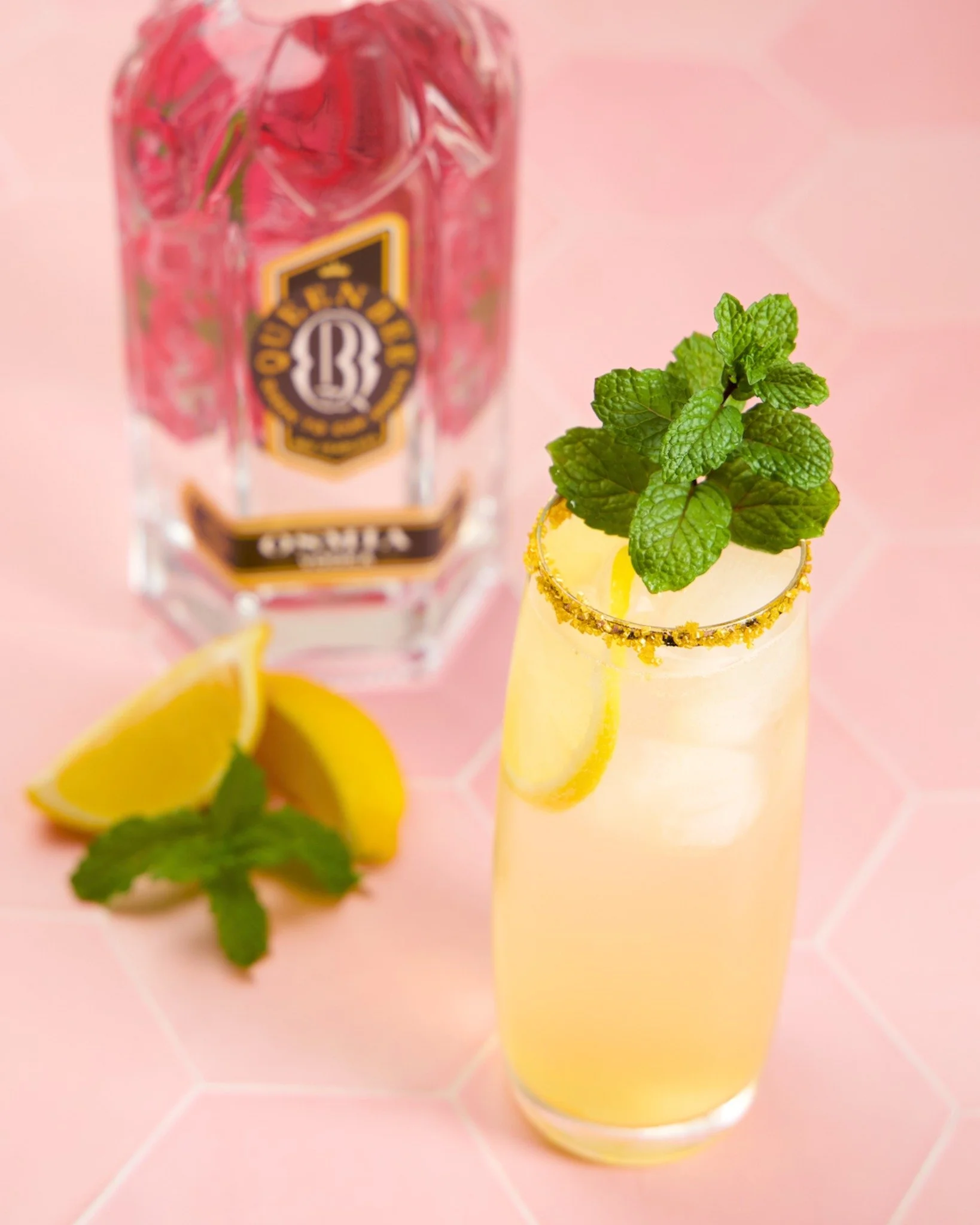 A tall glass of lemonade garnished with mint leaves and lemon slices on a pink tiled surface, with a bottle of vodka, lemon wedges, and mint sprigs nearby.