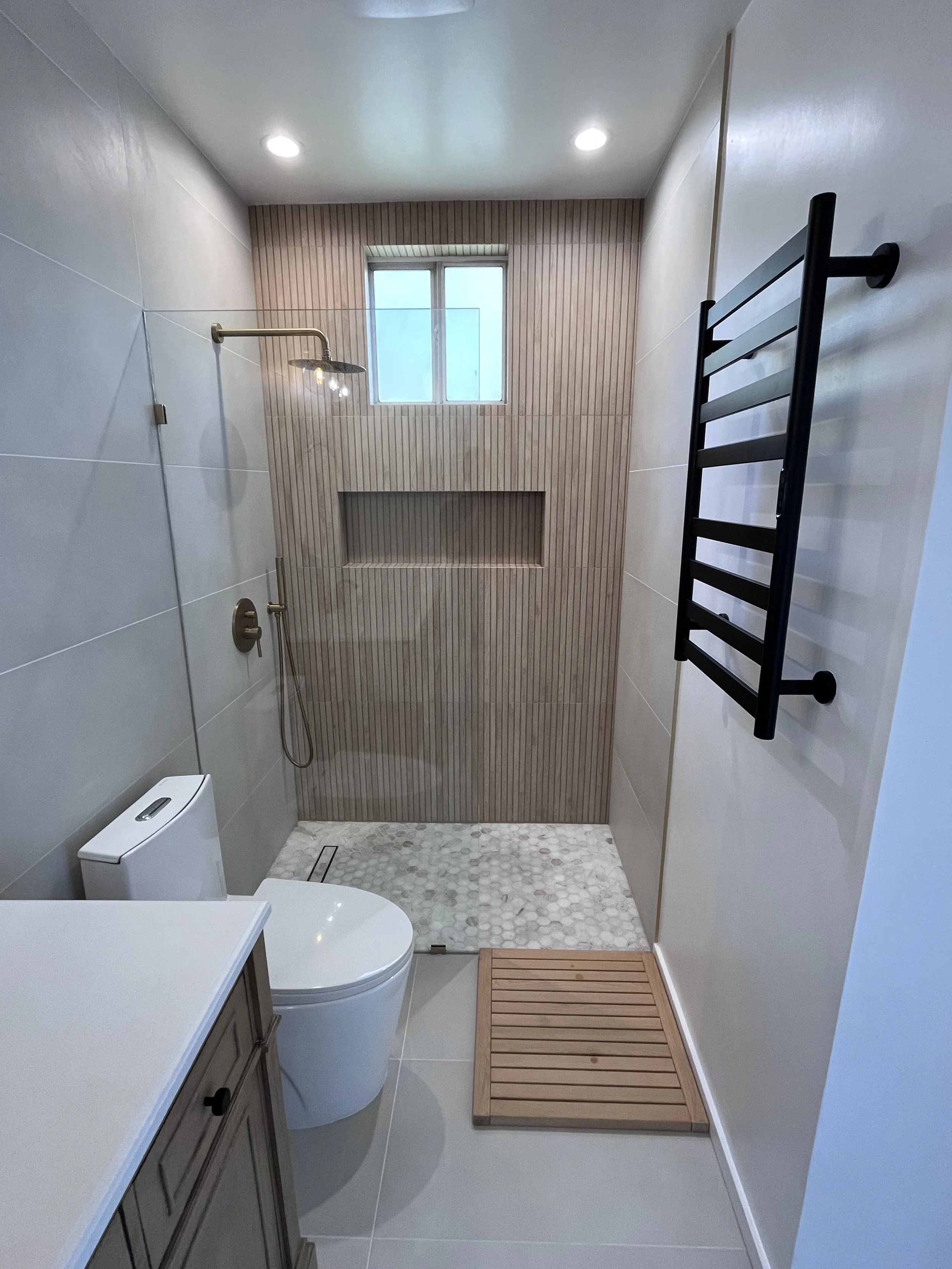 A modern bathroom with a walk-in shower, small window, black towel warmer, toilet, and a wooden bath mat.