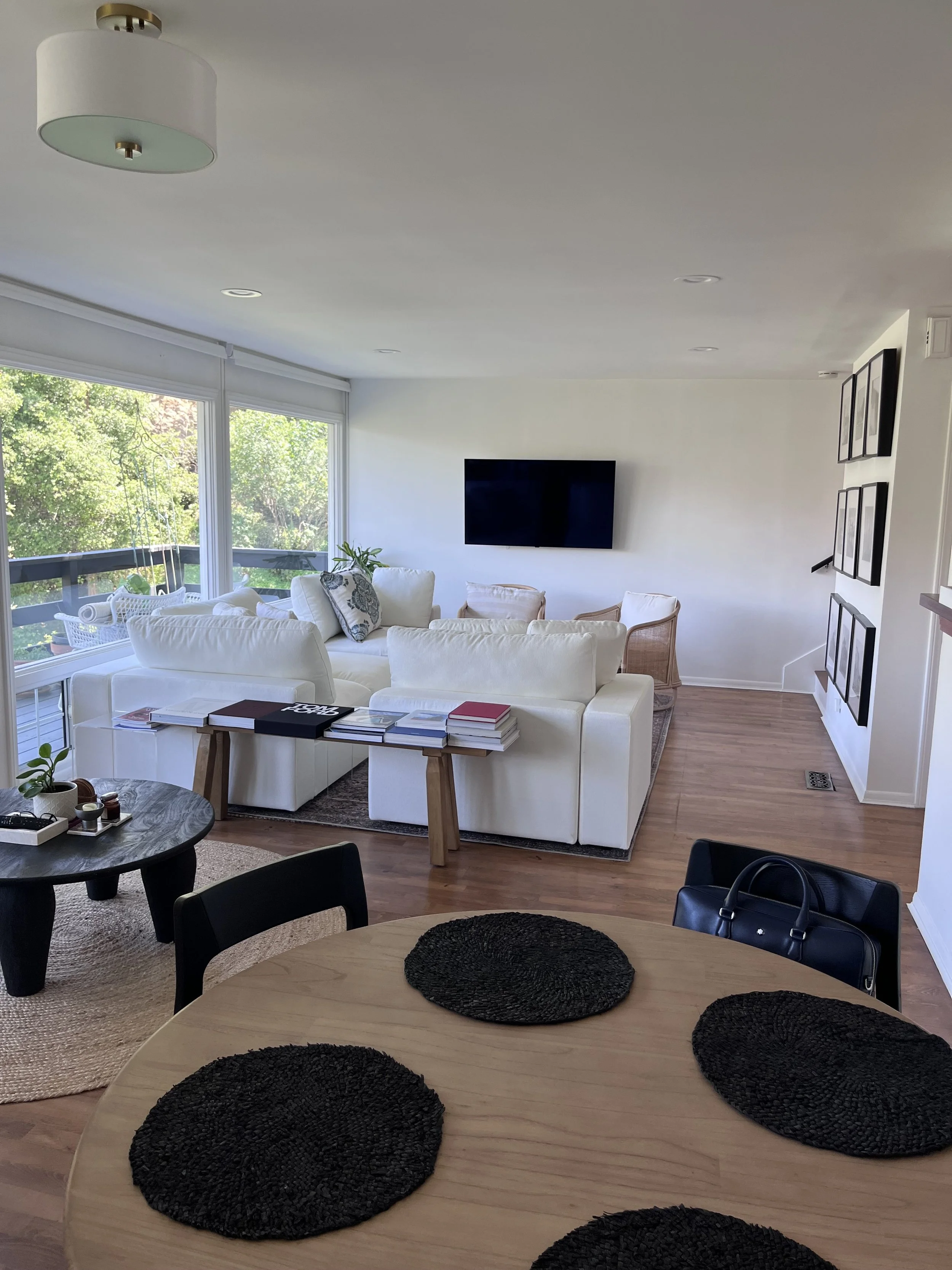 Living room with white sofa, black TV mounted on wall, glass windows with trees outside, wooden dining table with four black placemats, black chairs, and a small round black side table.