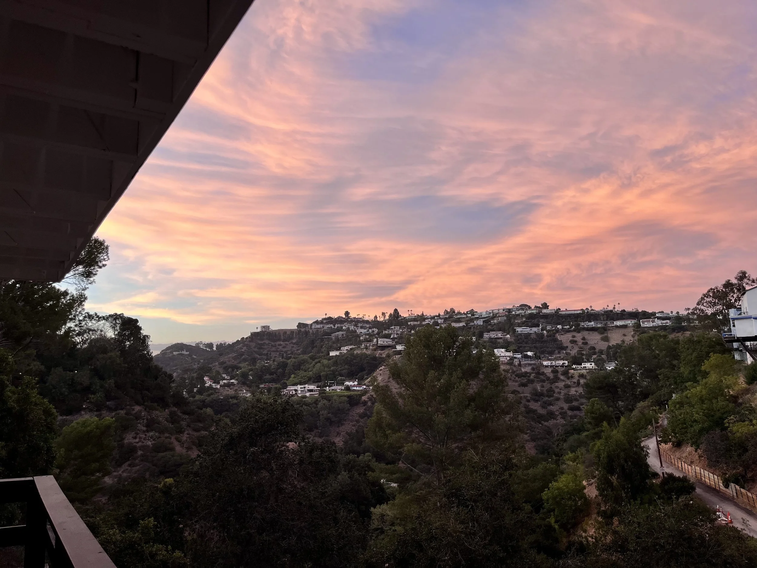 Sunset sky over a hillside residential area with trees and houses, seen from a balcony or porch.