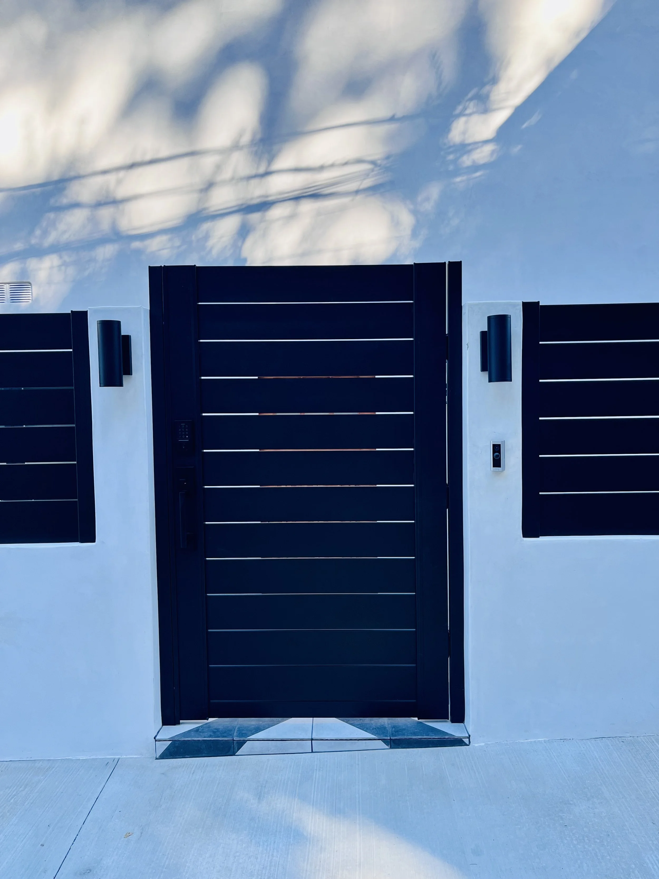 A modern black metal gate with horizontal slats, mounted on a white concrete wall, with a pathway in front and a blue sky with scattered clouds overhead.