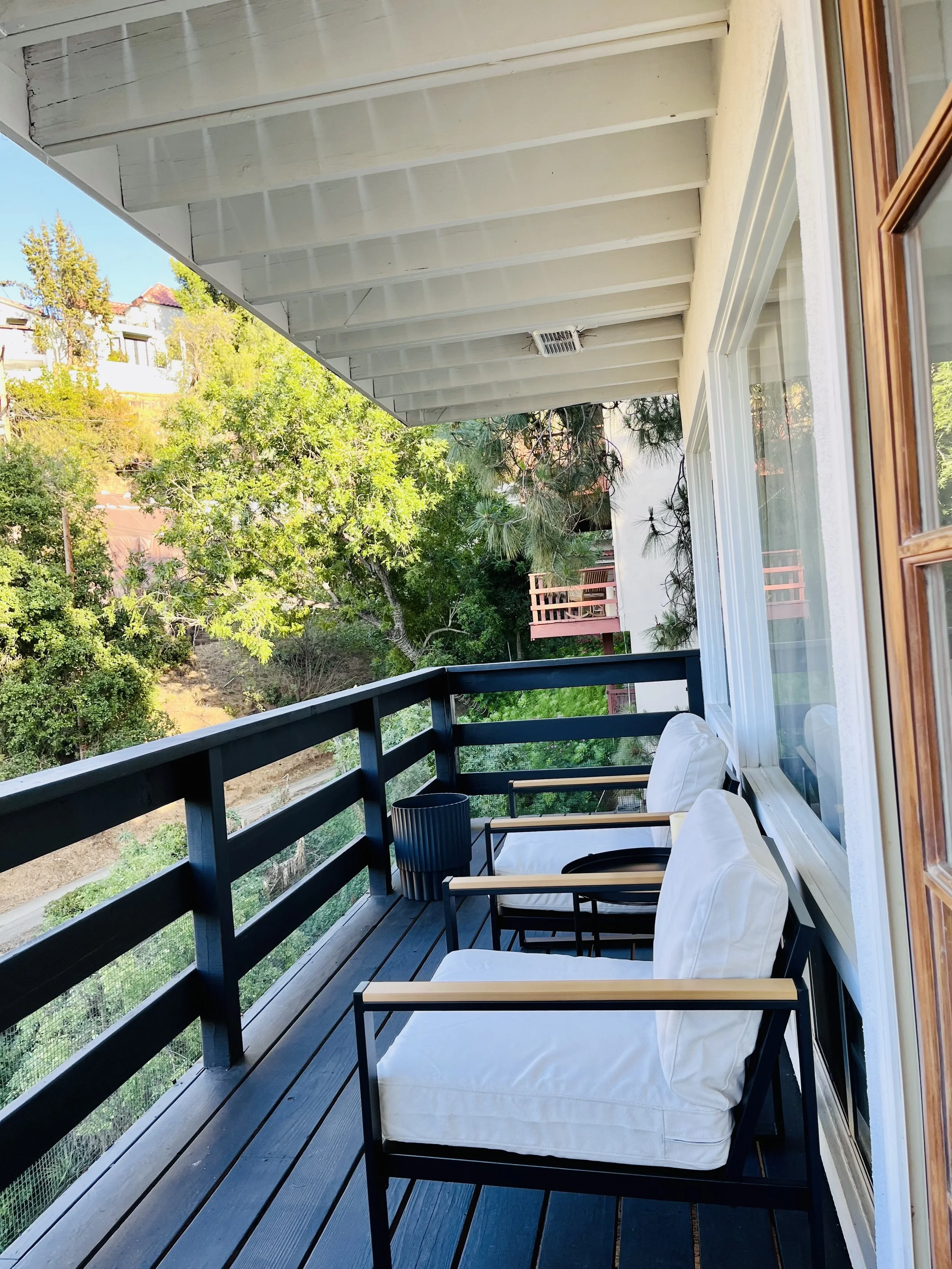 Balcony with black railing, wooden floor, and two black chairs with white cushions overlooking trees and neighboring houses.