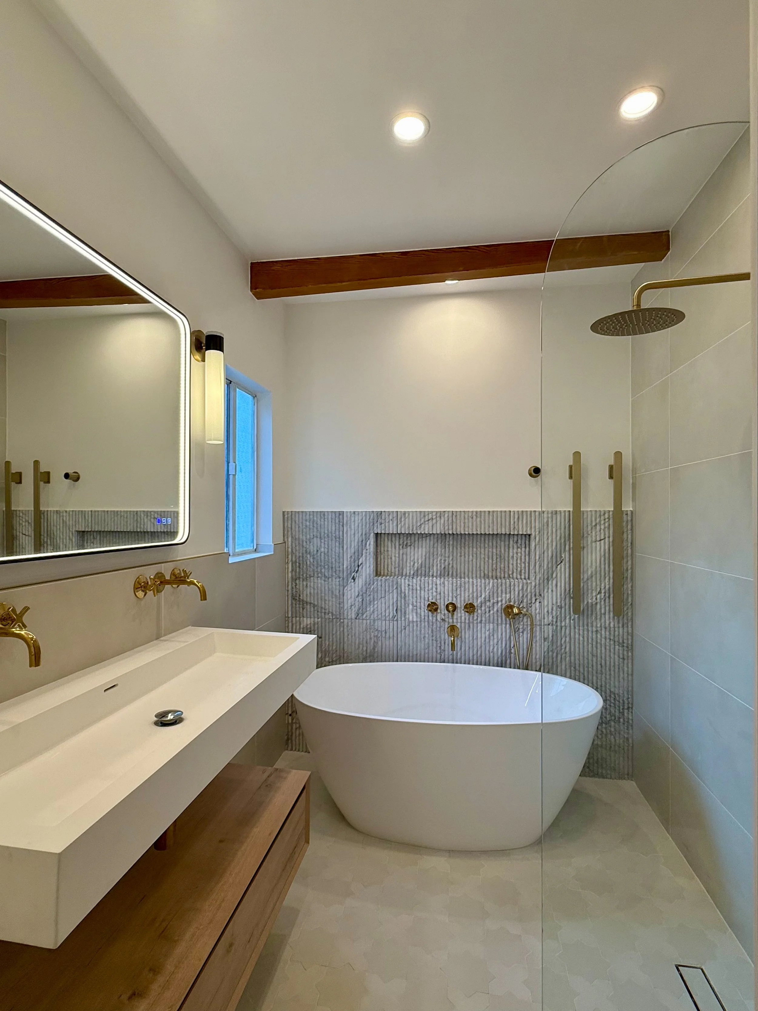 Modern bathroom featuring a large bathtub with brass fixtures, a rectangular white vanity with brass taps, a large illuminated mirror, a wall-mounted shower head, a small window, and wooden beams on the ceiling.