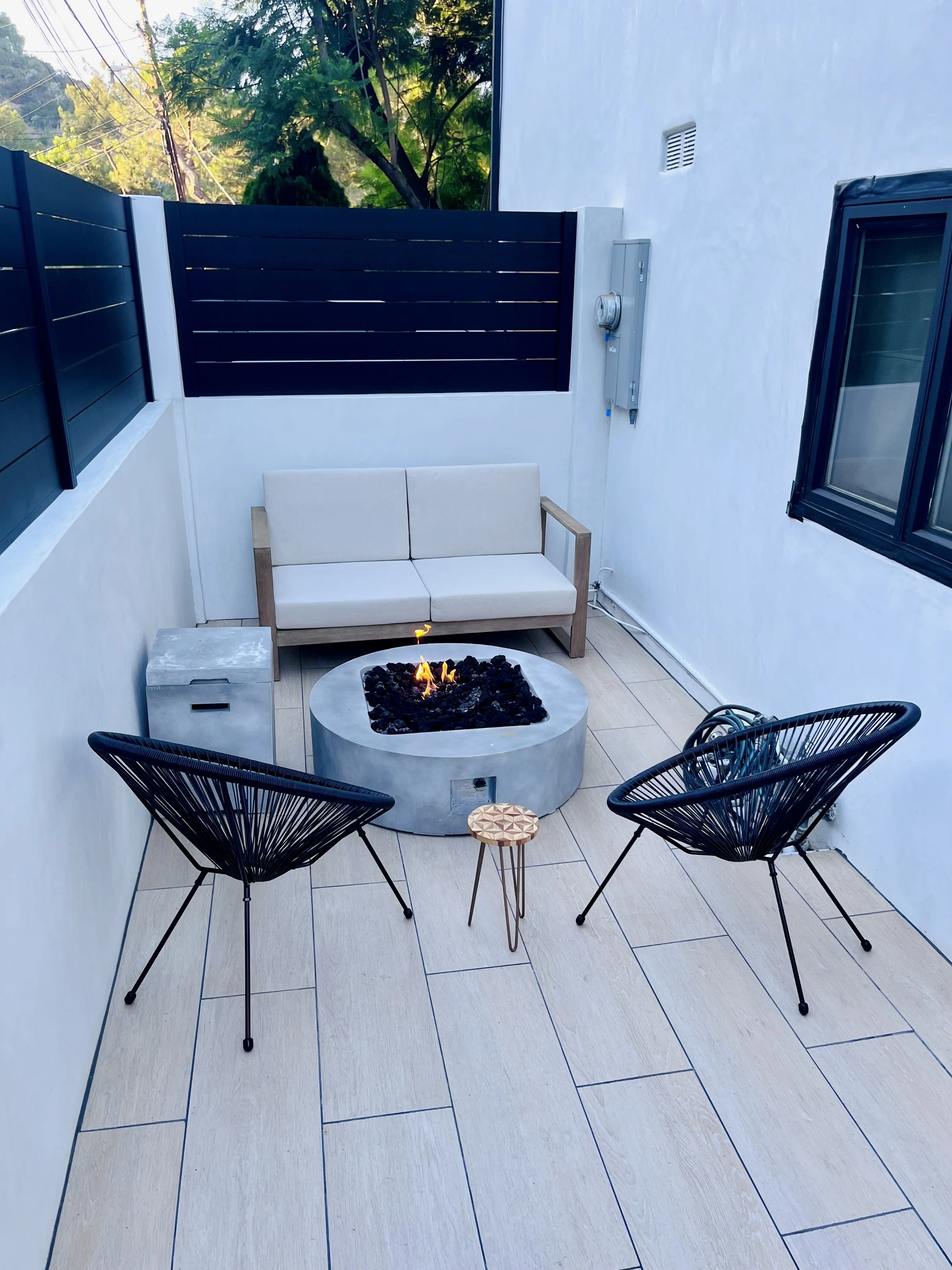 Small outdoor patio with two black wire chairs, a small round wooden side table, a white sofa, a circular fire pit with flames, and a white wall with a window and electrical panel.