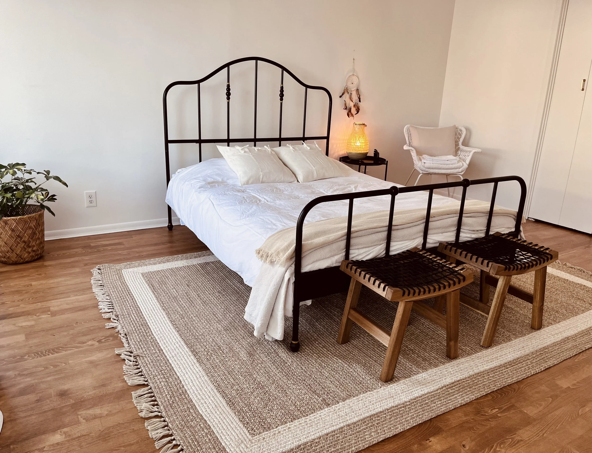 A cozy bedroom with a black metal bed frame, white bedding, and beige pillows. There are two wooden stools at the foot of the bed, a woven rug, a potted plant, a small black side table with a glowing yellow lamp, a white chair, and a dreamcatcher han