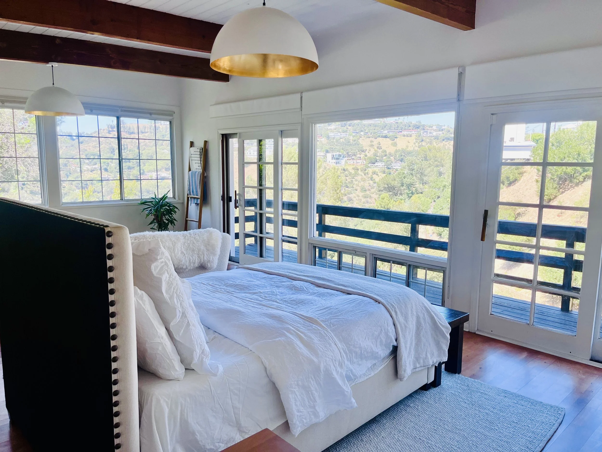 Bright bedroom with a large bed, white bedding, and a cream headboard with nailhead trim. Two large pendant lights with white and gold accents hang from the ceiling. Floor-to-ceiling windows and a sliding glass door open to a balcony with a view of a