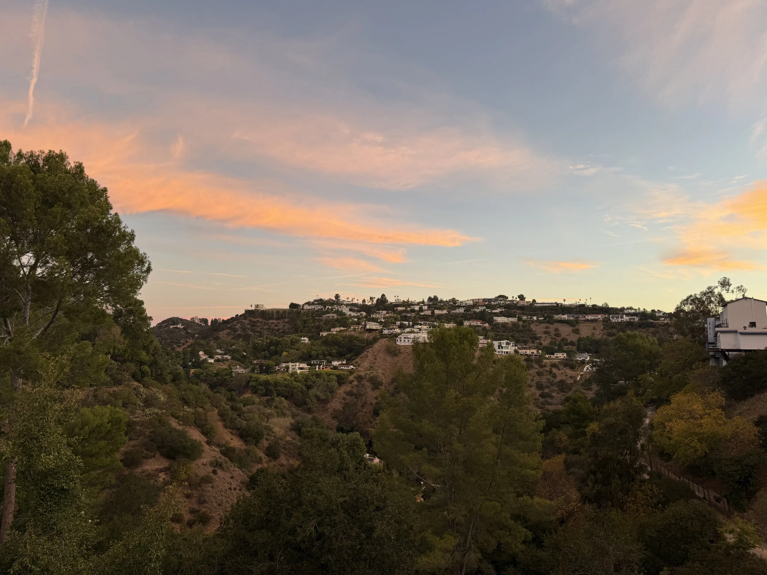 Sunset view of a hillside with scattered houses, trees, and a colorful sky with pink and orange clouds.