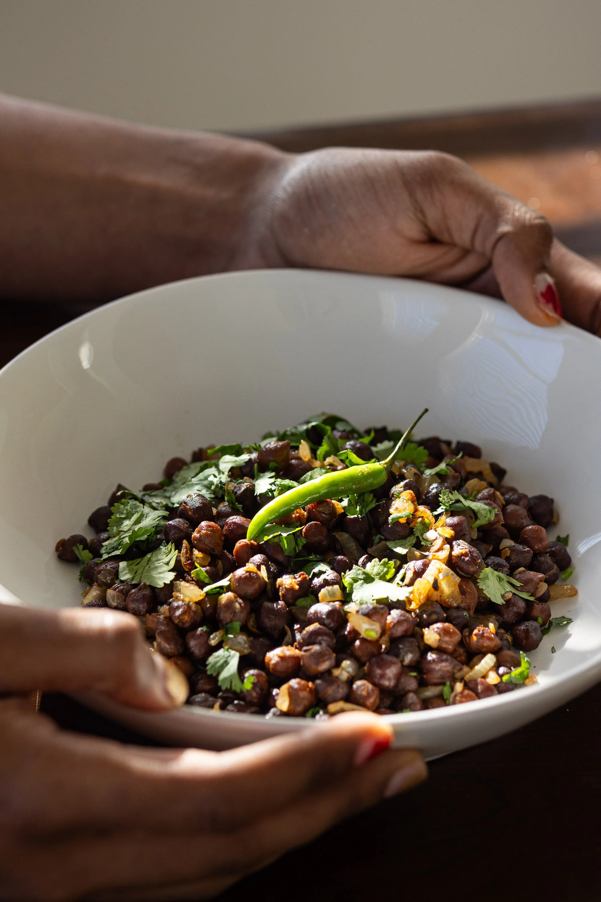 A person holding a white bowl filled with cooked black chickpeas, garnished with chopped herbs, a green chili, and chopped onions.