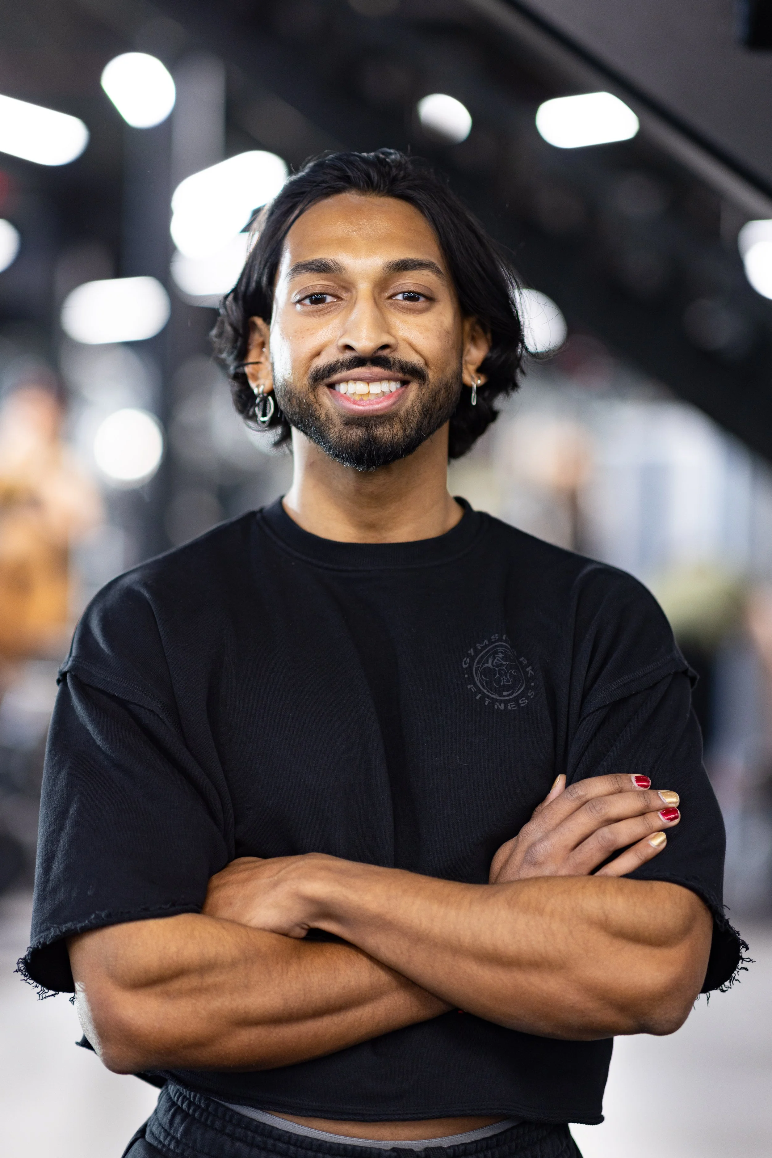A young man with black hair, a beard, and earrings, smiling with arms crossed, in a gym setting.