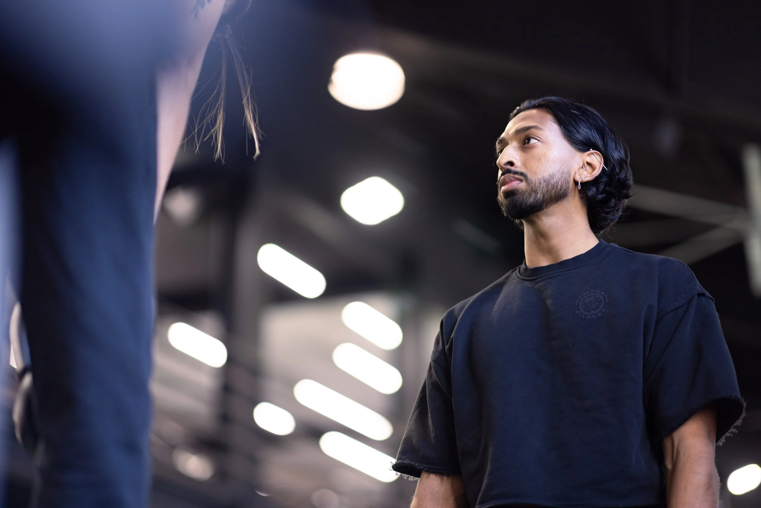 A young man with dark, wavy hair, beard, and earrings, looking attentively at another person in a black shirt, indoors with bright ceiling lights and dark ceiling panels.