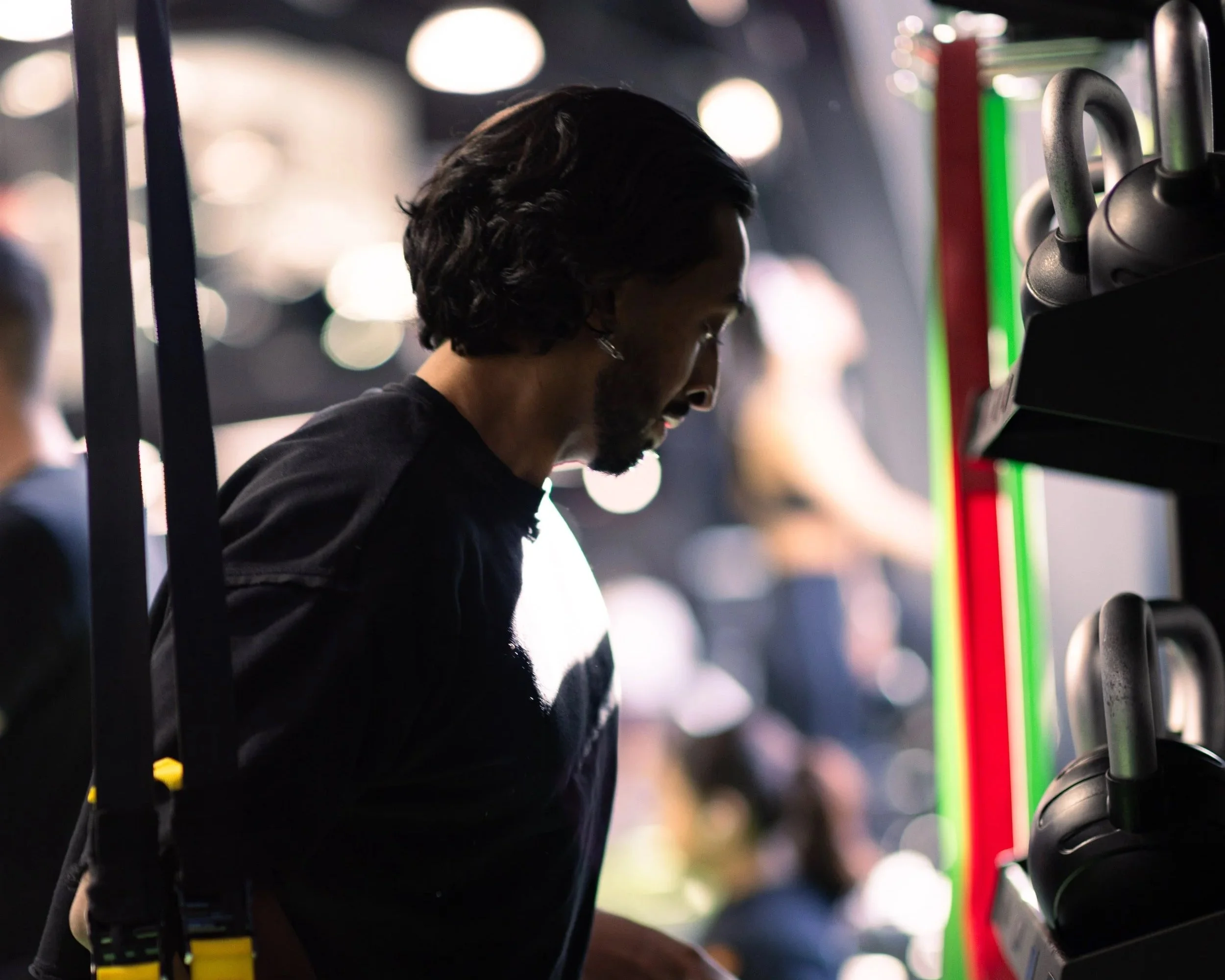 A man with long dark hair and a beard, wearing a black shirt, is working out in a gym with black kettlebells on a rack in front of him. The background is blurred with round lights and other gym-goers.