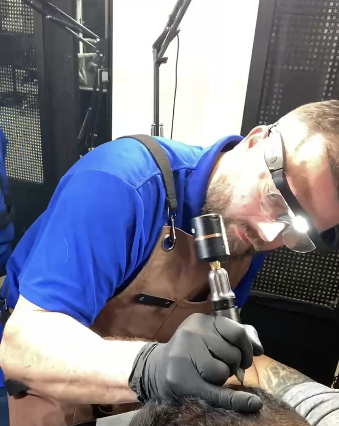 A scalp micropigmentation artist in a blue shirt, wearing safety glasses and gloves, working on a person's hair density treatment.