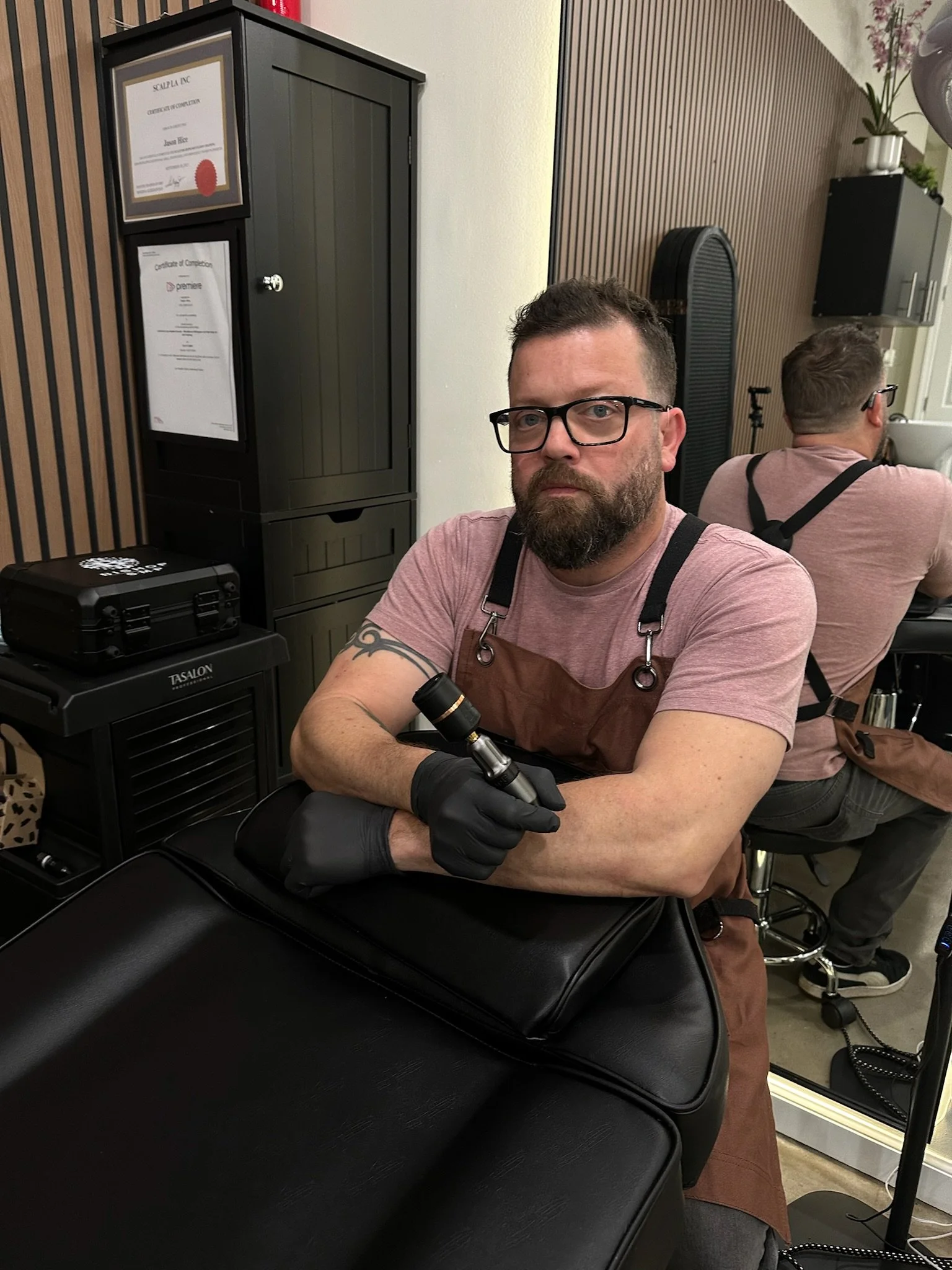 A man with glasses, a beard, and tattoos, wearing a pink t-shirt and brown apron, sitting at a SMP station holding an SMP machine, with another person in the background. The setting appears to be a scalp micro pigmentation studio.
