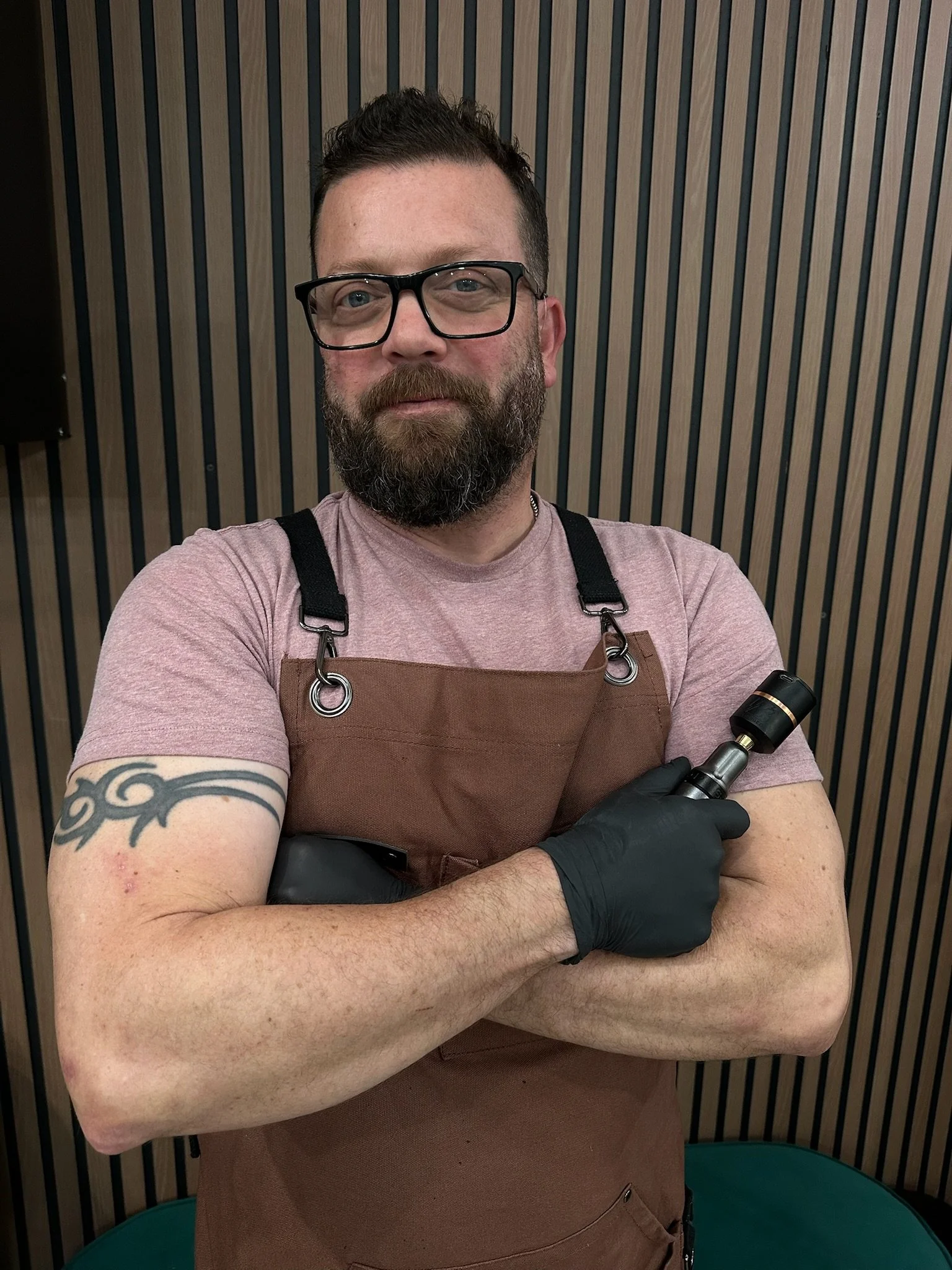 A man with glasses, a beard, and a tattoo on his arm wearing a brown apron and black gloves, holding a scalp micropigmentation machine, standing in front of a wood-paneled wall.