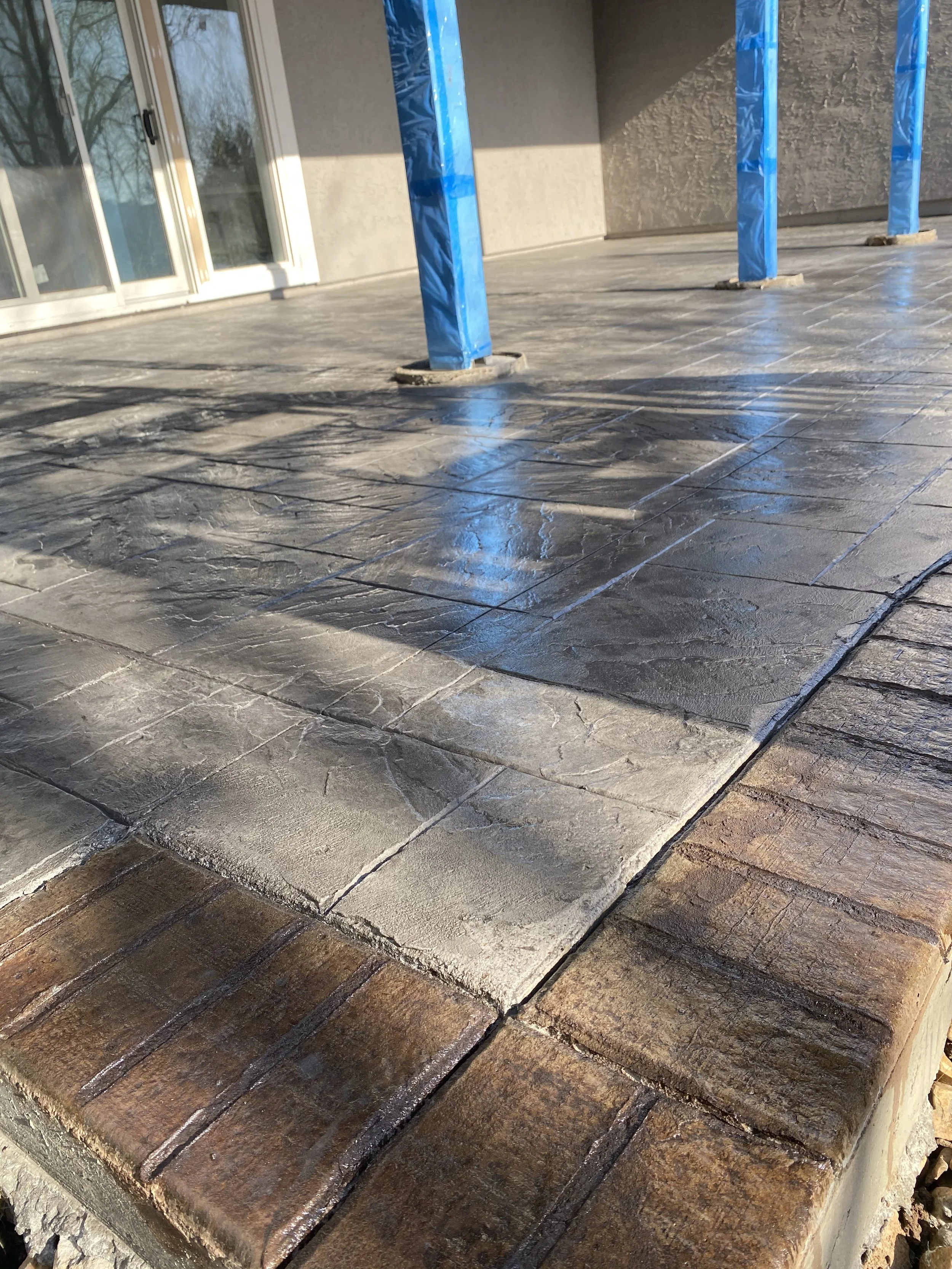 Newly installed stone patio with a textured surface, featuring a mix of natural stone colors and a slightly raised brick border, with blue protective wraps on post bases.