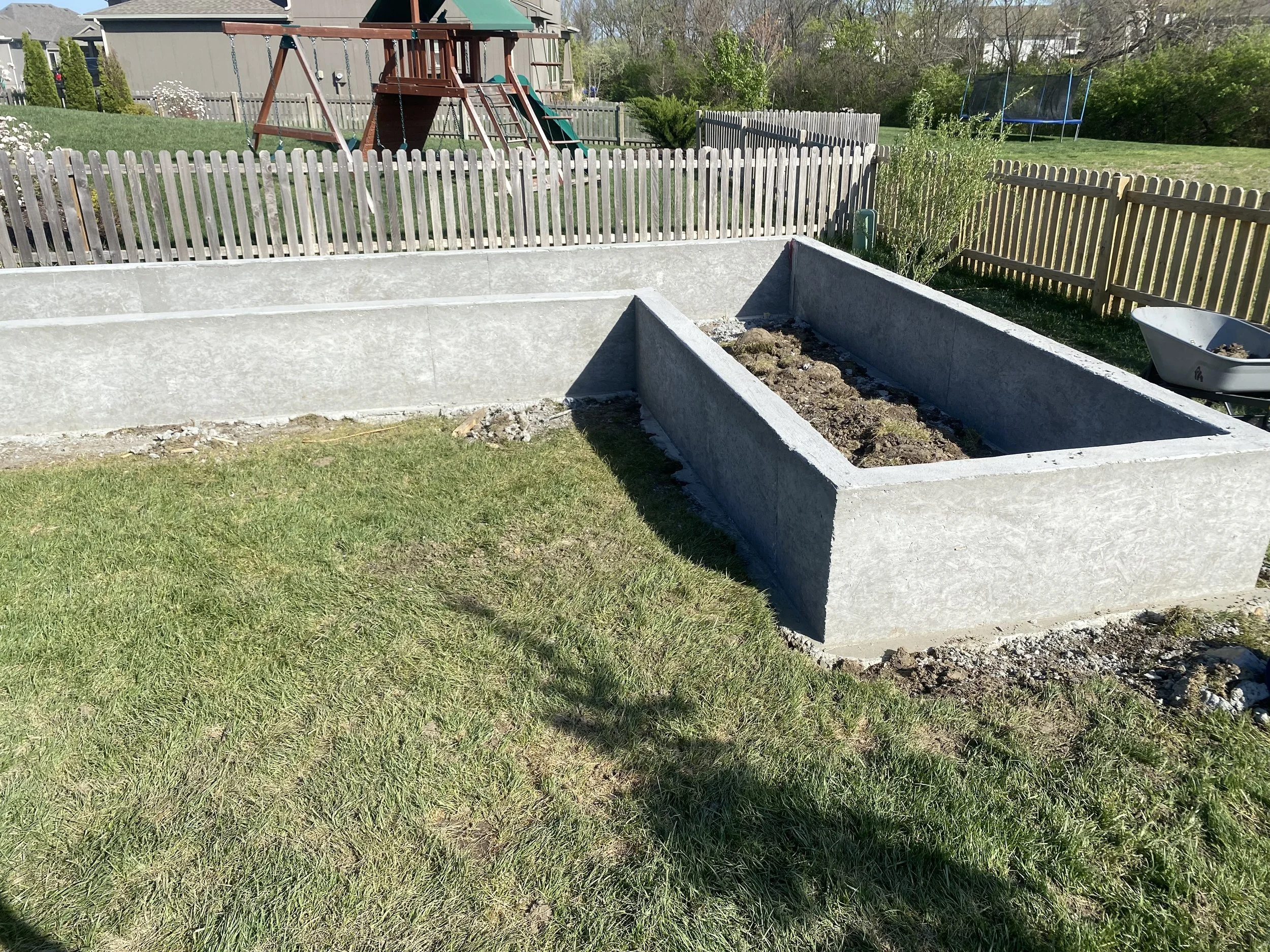 Garden beds with concrete borders, one is empty and the other has soil, in a backyard with a wooden fence, grass, a playset, and a trampoline.