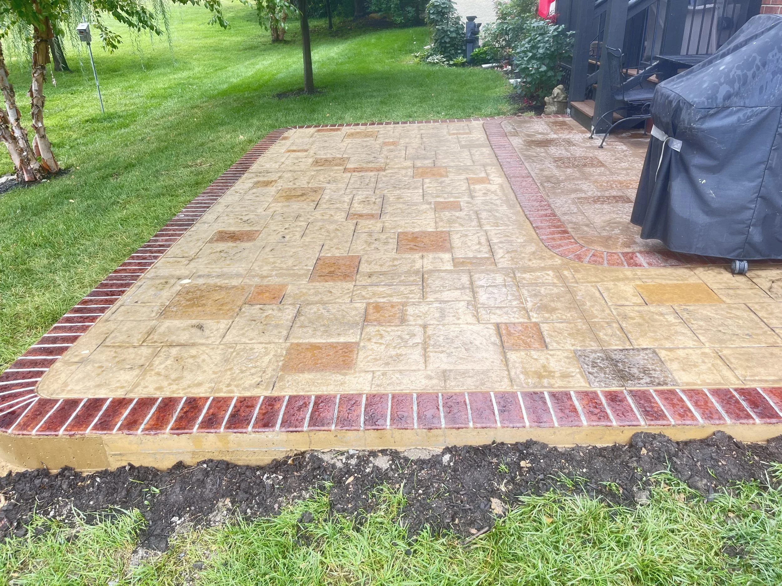 Newly paved brick and stone patio in a backyard with a grassy area, trees, and a large covered grill.