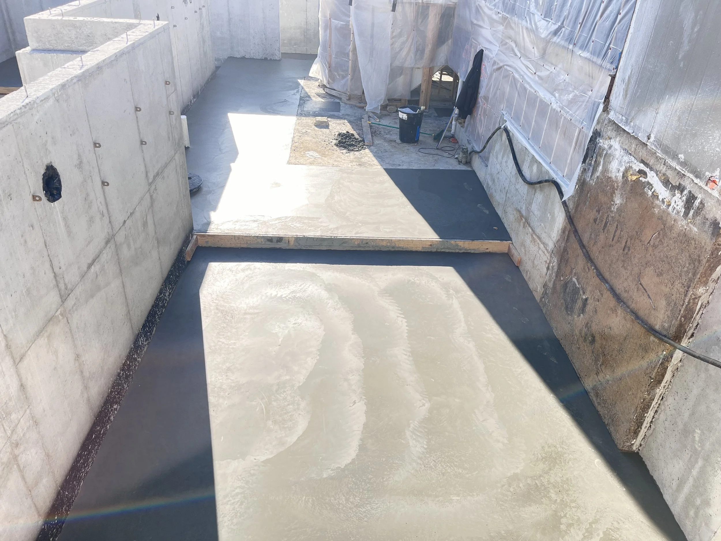 Construction site with freshly poured concrete floor, partially shaded, surrounded by unfinished walls and construction tools.