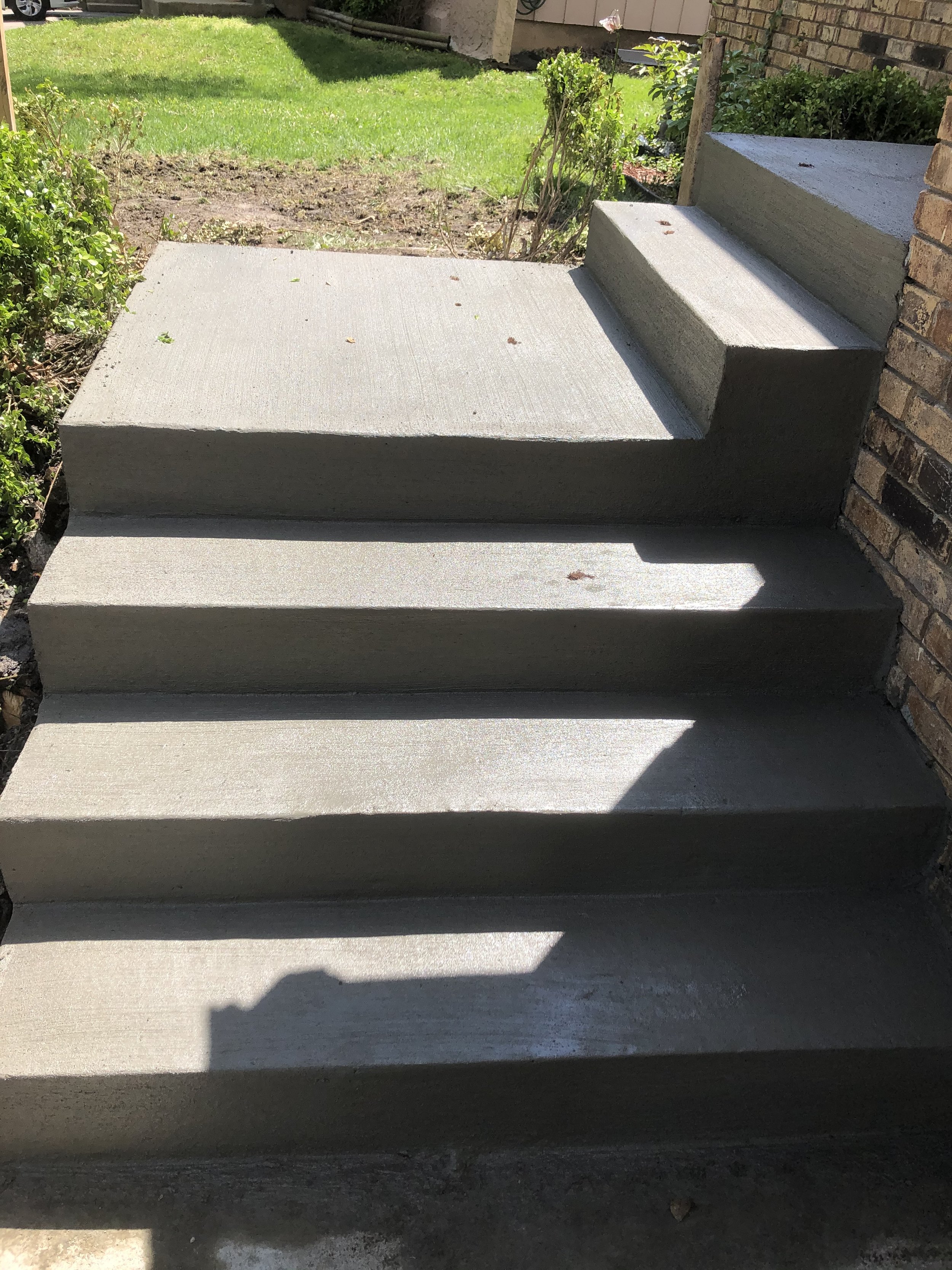 Concrete steps leading up to a porch, with shadows cast by the sunlight, bordered by a brick wall on the right and a garden area with grass and small plants on the left.