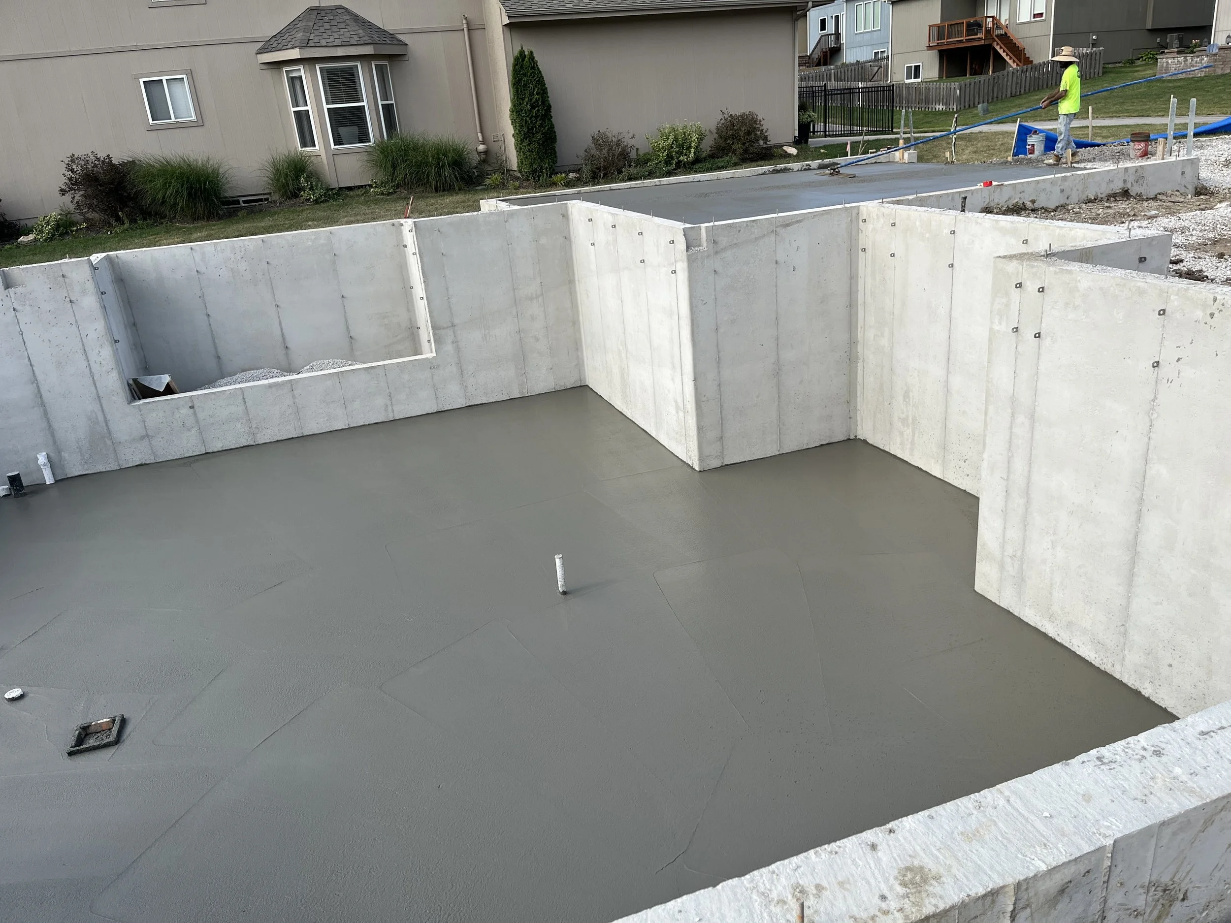 Foundation of a building under construction with concrete sump and walls. Worker in bright yellow shirt and hat working nearby.