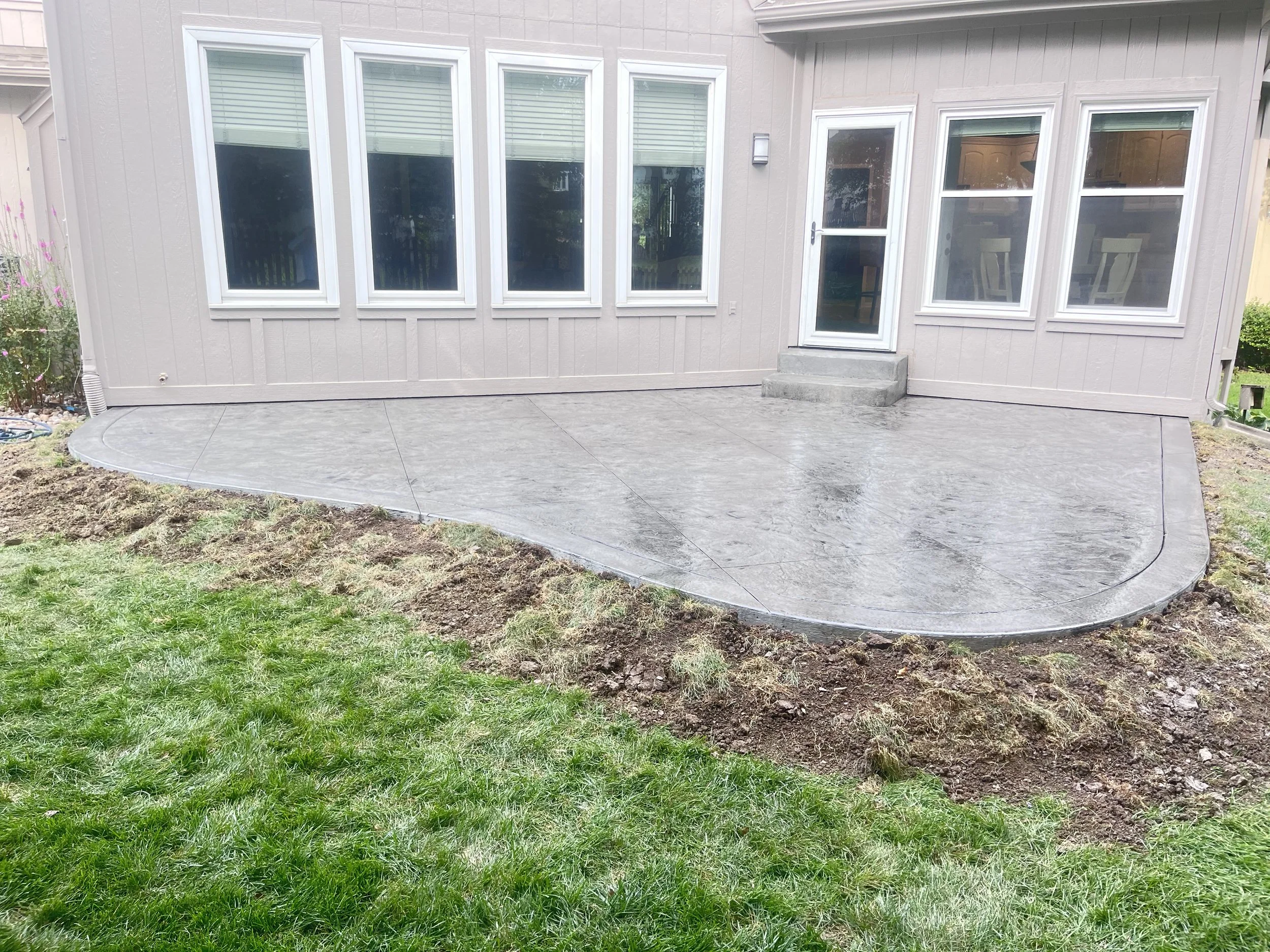 Newly poured concrete patio with a curved edge, adjacent to a house with multiple windows and a door, in a backyard with grass and some soil.