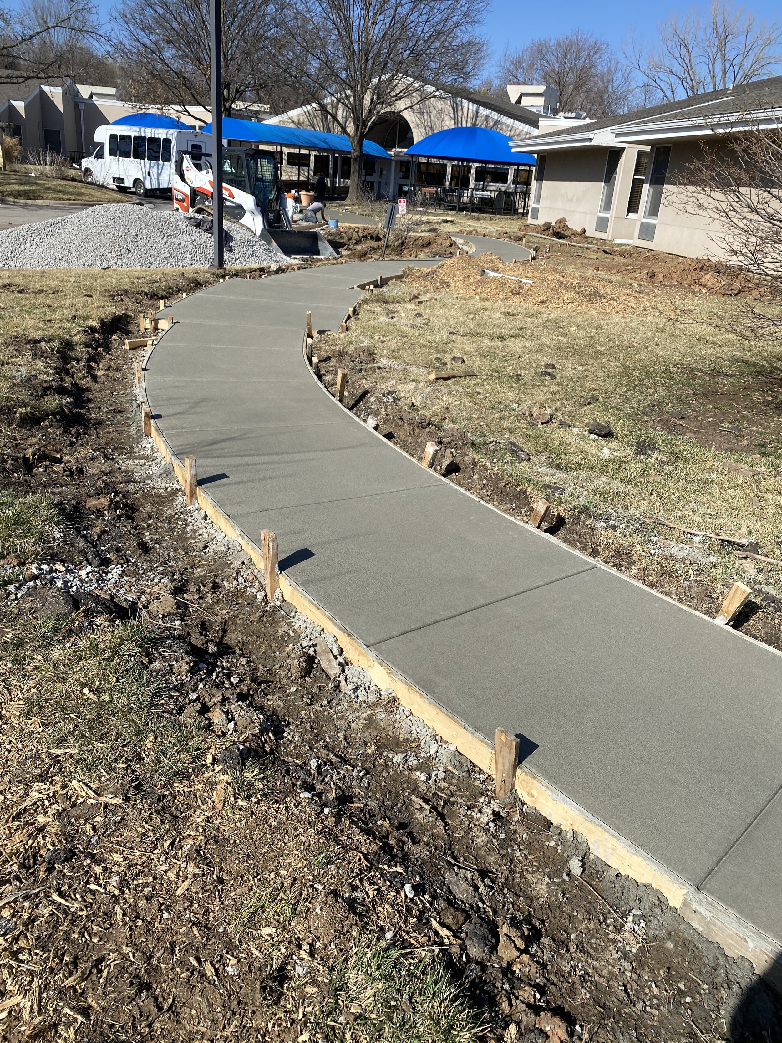 Newly poured concrete sidewalk winding through a grassy yard with construction stakes along the edges, under a clear blue sky.