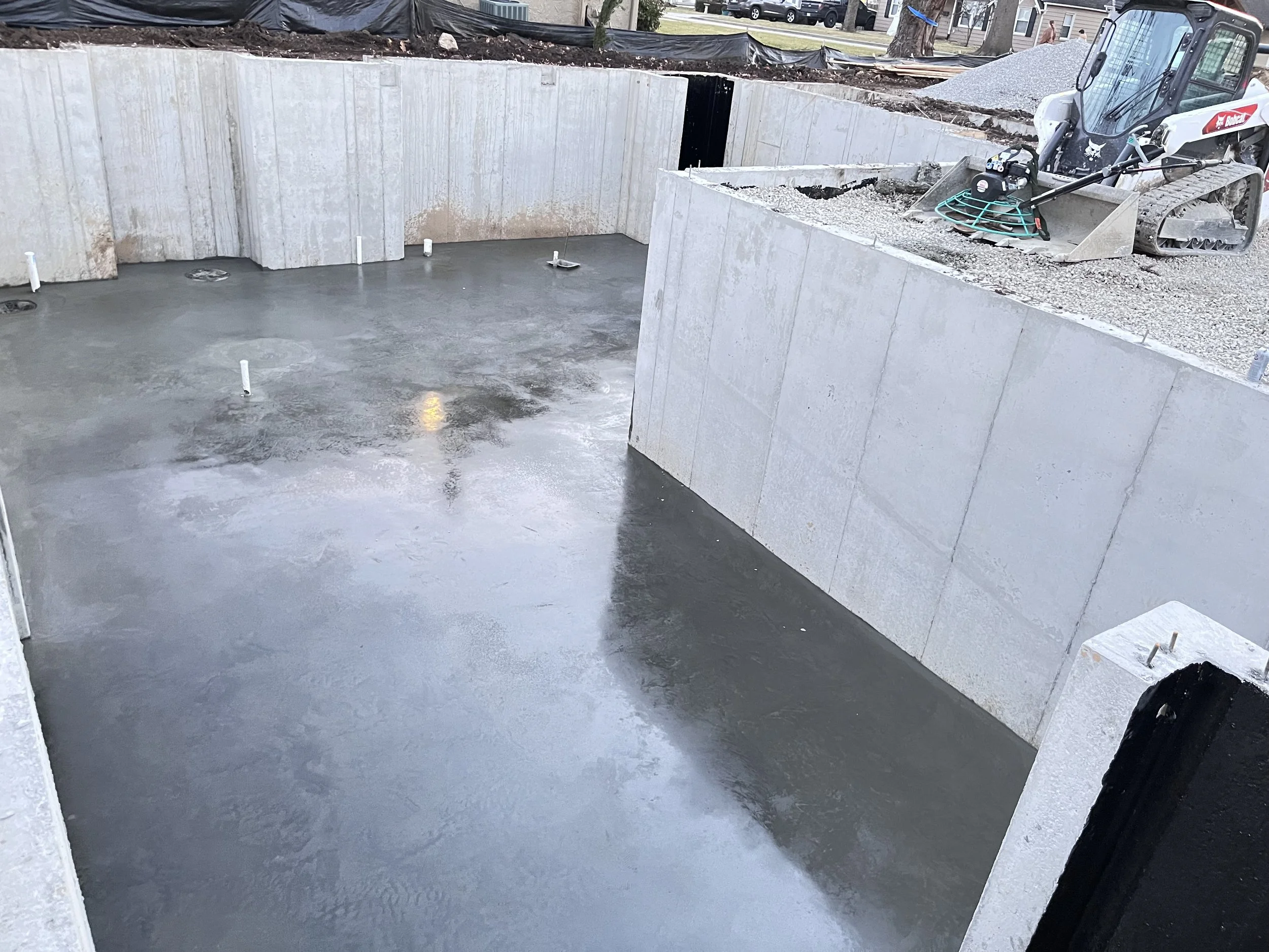 Freshly poured concrete in a building foundation with plumbing pipes protruding, with construction equipment and gravel visible at the site.