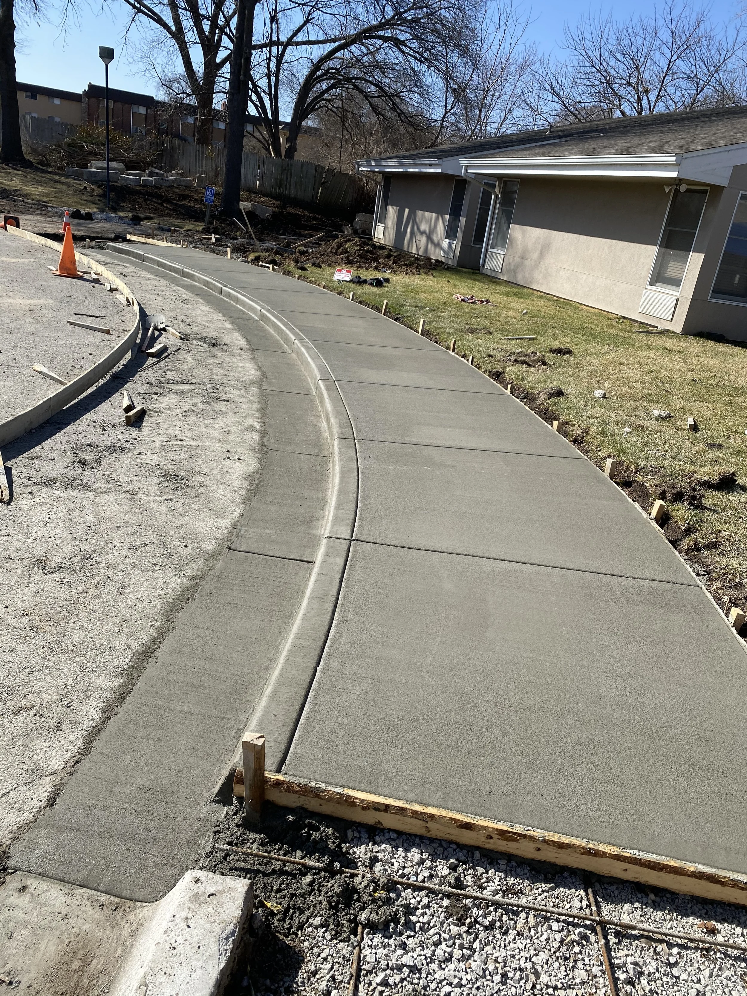 Newly poured concrete sidewalk curved along a residential lawn with construction tools and warning cones nearby during daytime.